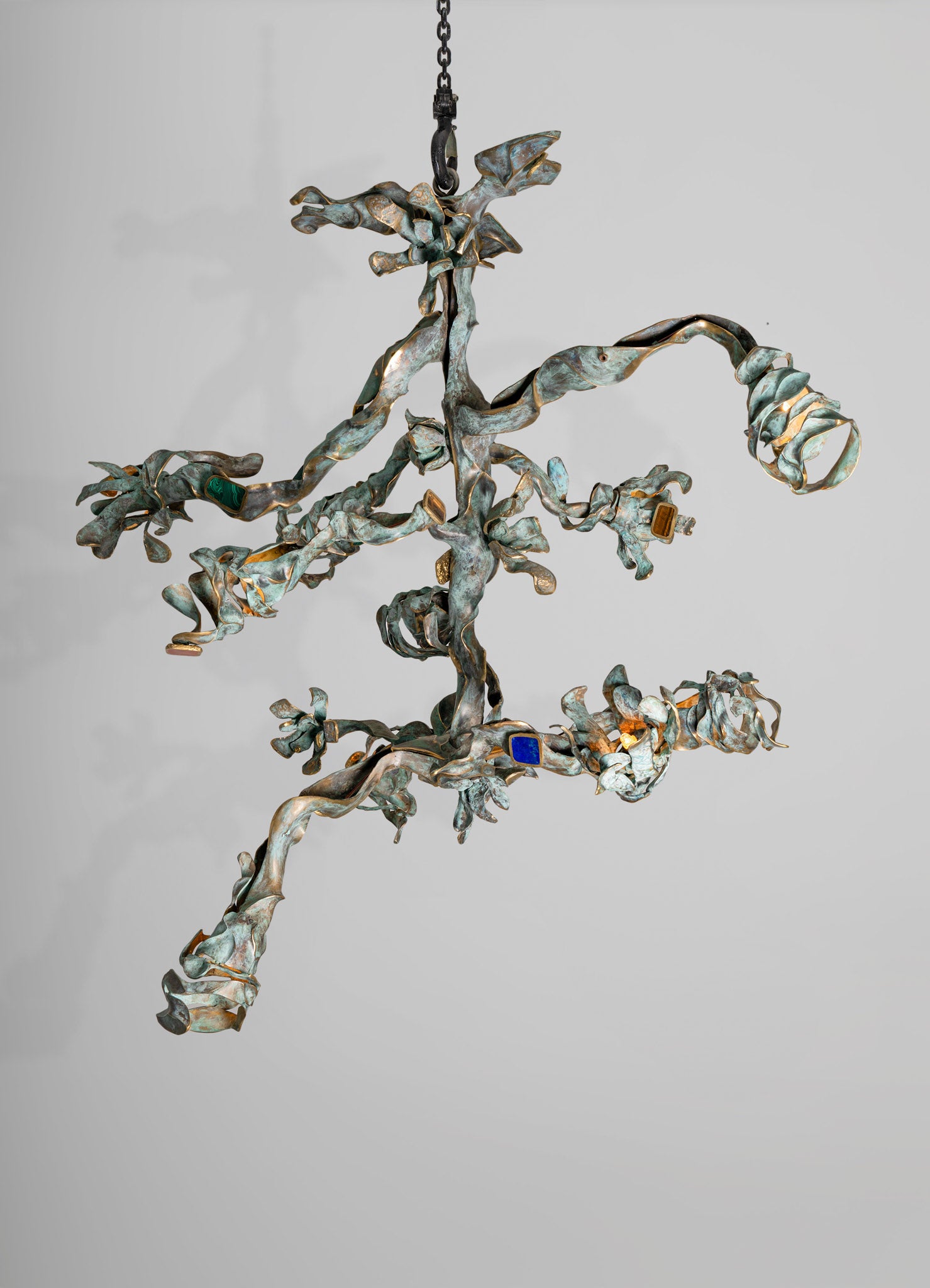 A sculptural chandelier made of twisted metal with an aged bronze finish, featuring small colored glass accents in blue, green, and amber, hanging against a plain background.