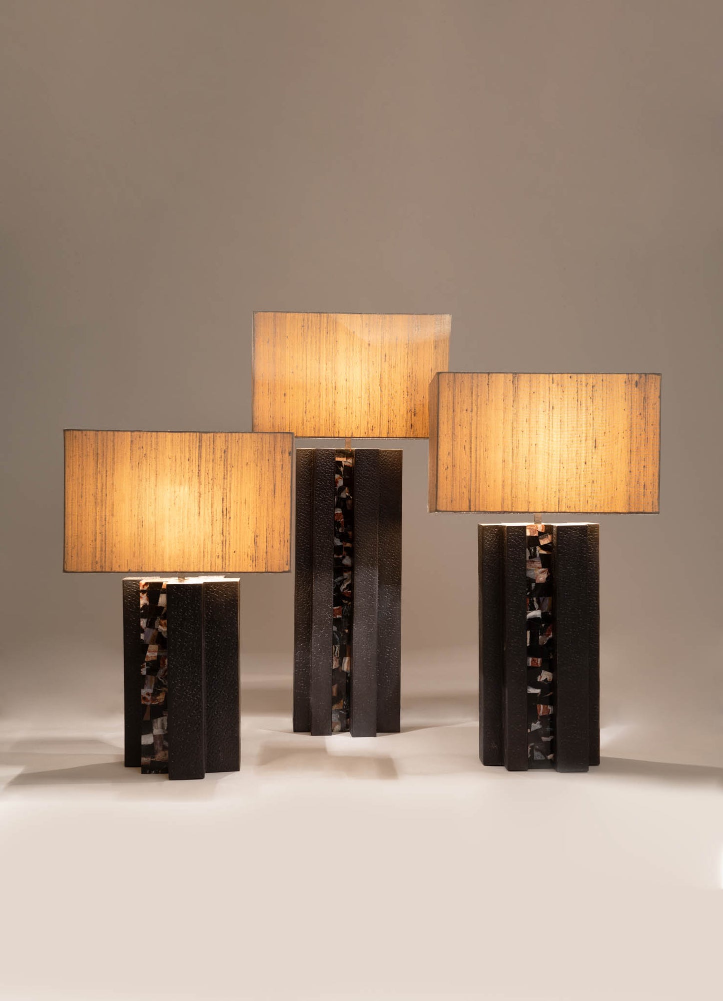 Three table lamps with rectangular fabric shades in warm beige tones, mounted on black bases with textured and marbled details, arranged on a neutral surface. The lamps have a contemporary design with a blend of modern materials and clean lines.