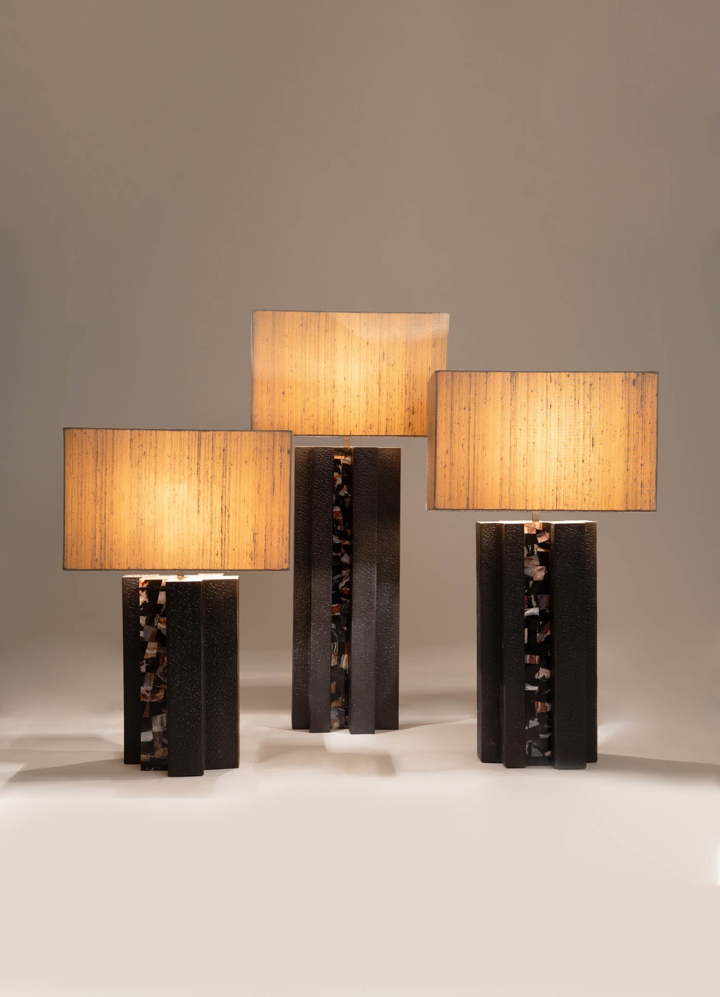 Three table lamps with rectangular fabric shades in warm beige tones, mounted on black bases with textured and marbled details, arranged on a neutral surface. The lamps have a contemporary design with a blend of modern materials and clean lines.