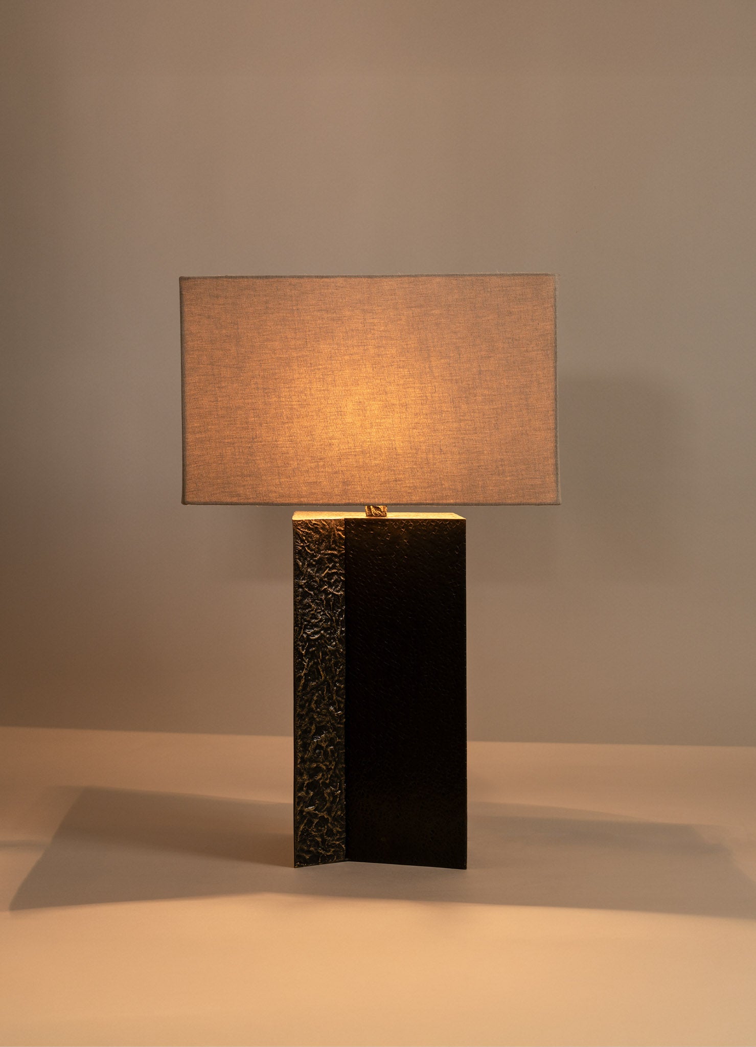 A table lamp with a rectangular beige fabric lampshade and a cylindrical base crafted from textured black and gold metal, creating a modern and sophisticated aesthetic.