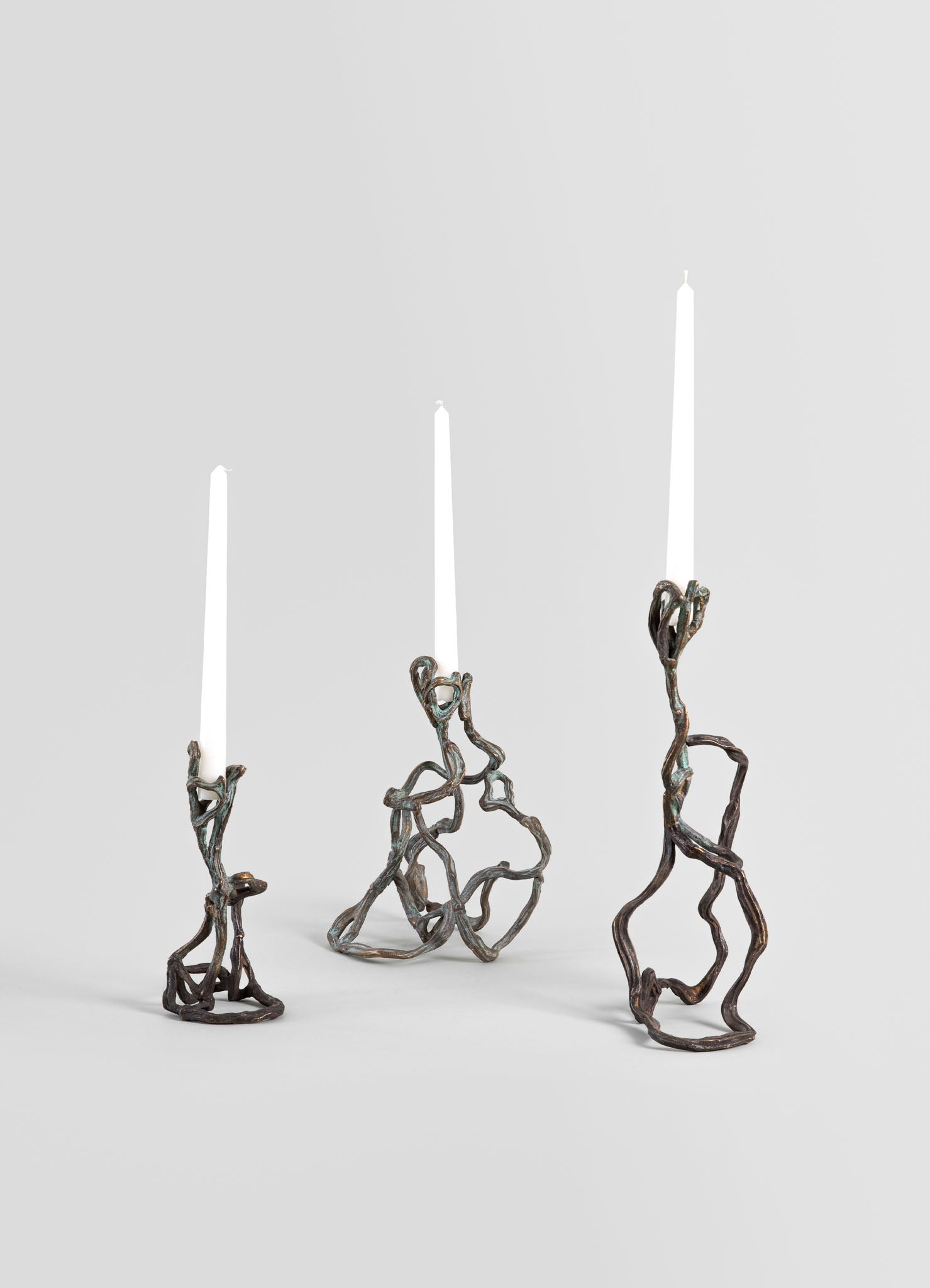 Three candlesticks made of twisted, branch-like metal with a dark bronze finish hold slender white candles. The design is organic and sculptural, with a rustic and contemporary style.