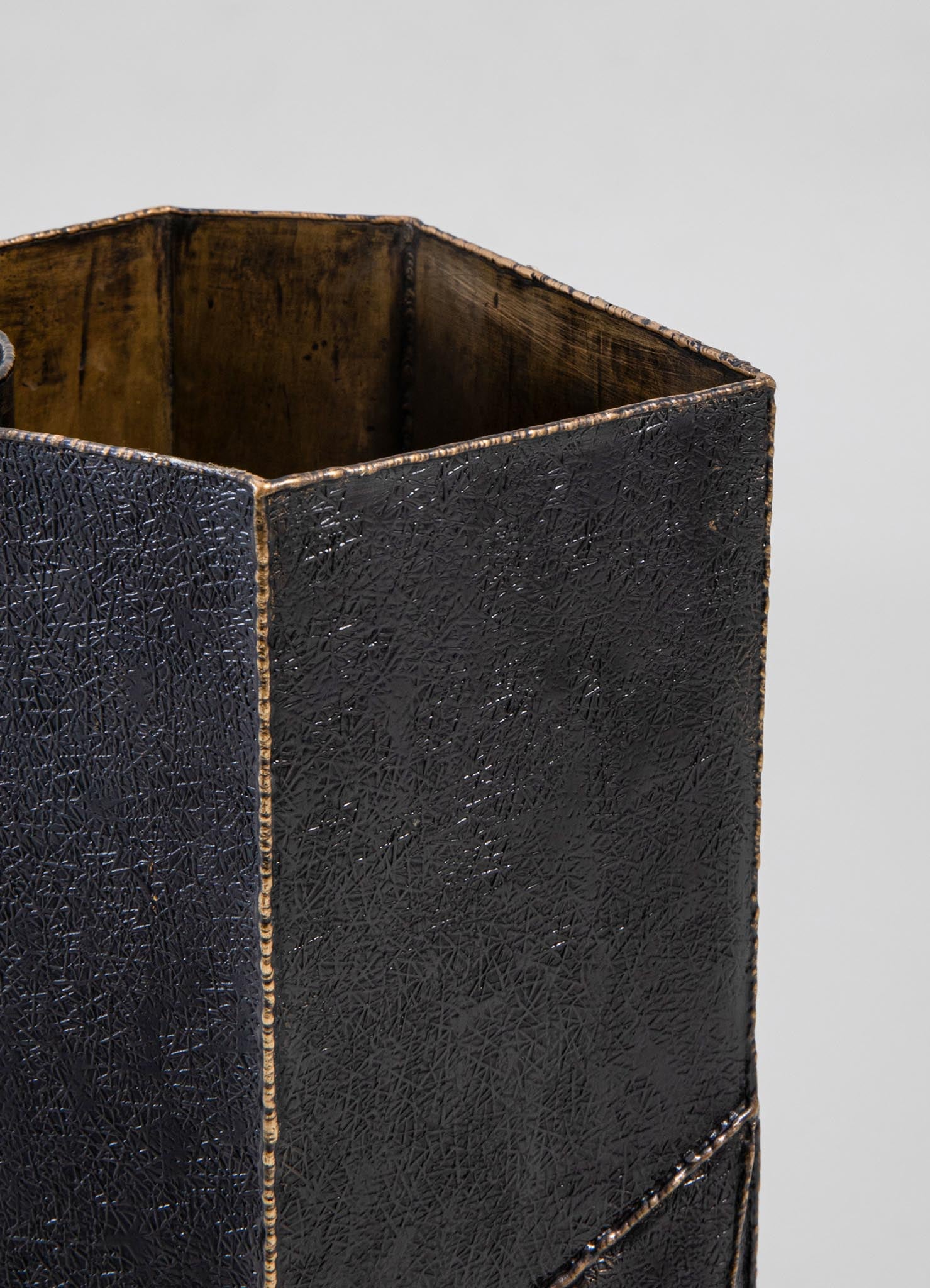 A black textured metal container with a geometric, crisscross pattern on its surface, featuring visible inside surfaces with a warm brown finish and subtle gold accents along the edges.