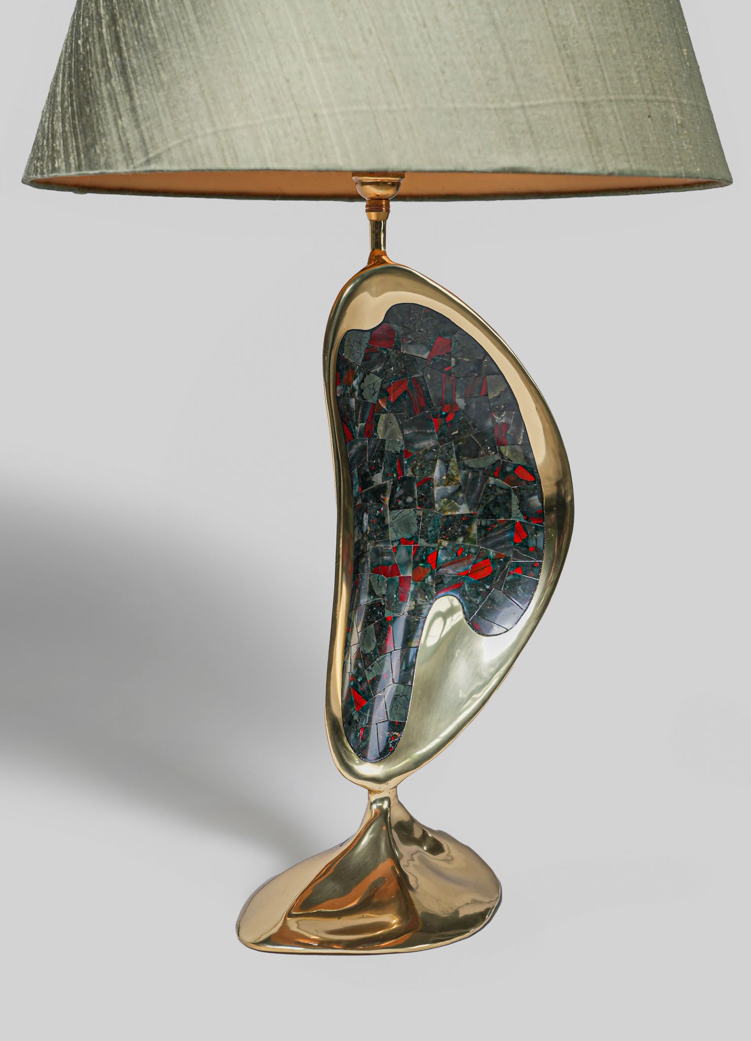 A table lamp featuring a gold-toned metallic base with an abstract, irregular shape and inlaid with multicolored mosaic stones in red, black, and gray. The lamp has a large, textured fabric shade in a muted green or taupe tone, creating a contemporary and artistic style.