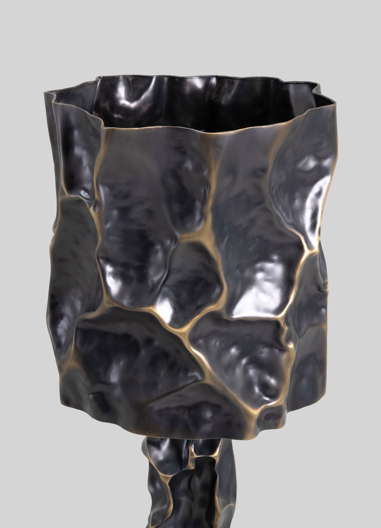 A decorative vase made of metal with a hammered, irregular surface in a dark, oxidized finish, featuring subtle gold accents along the creases. It has a sculptural, organic form with a textured, contemporary design.