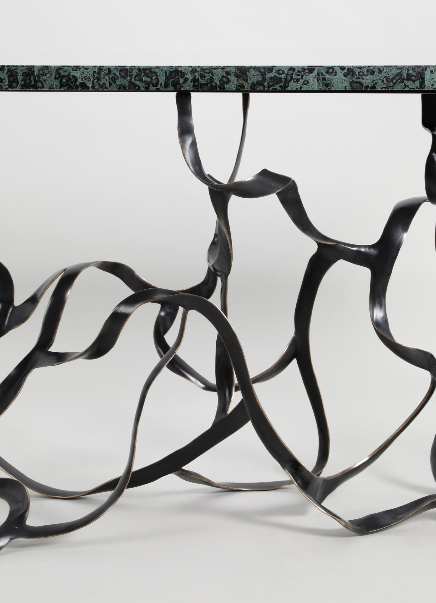 A table with a top surface made of dark green marble with black and beige veining, supported by a sculptural black metal base with twisted, organic forms.