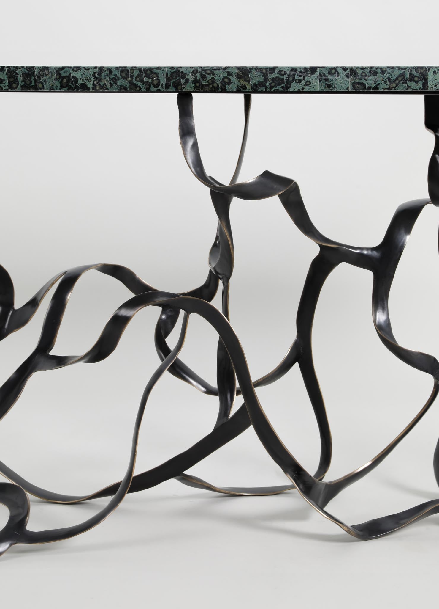 A table with a top surface made of dark green marble with black and beige veining, supported by a sculptural black metal base with twisted, organic forms.