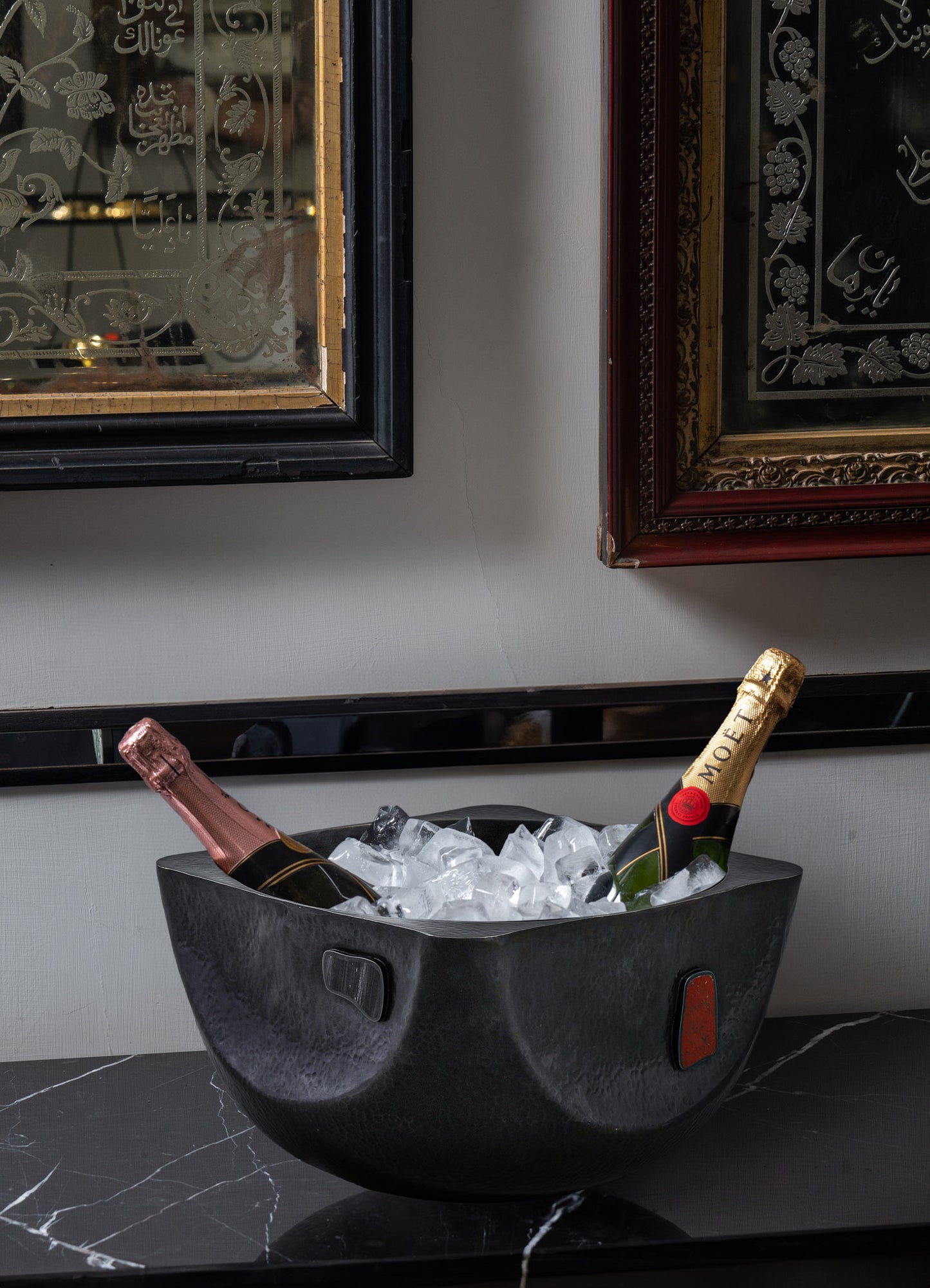 A black textured ice bucket with a rounded shape and small handles, holding two bottles of sparkling wine or champagne and ice cubes, placed on a dark marble surface. Framed artwork, mirrors, and decorative frames are visible in the background.