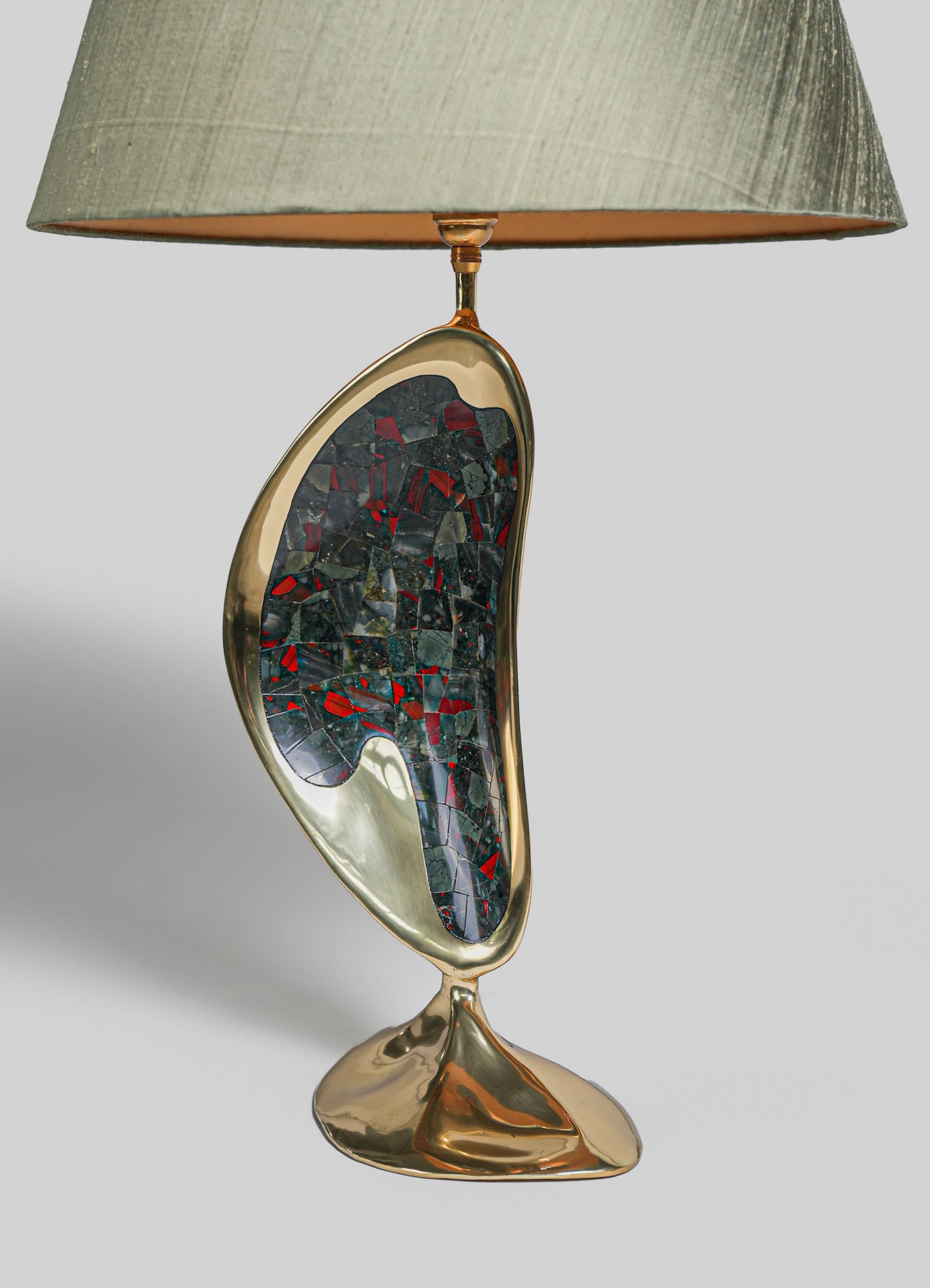 A table lamp featuring a carved, crescent-shaped base made of shiny brass with a mosaic inlay of black and red stones. It has a large, neutral beige fabric shade with a textured finish, set against a plain light gray background.