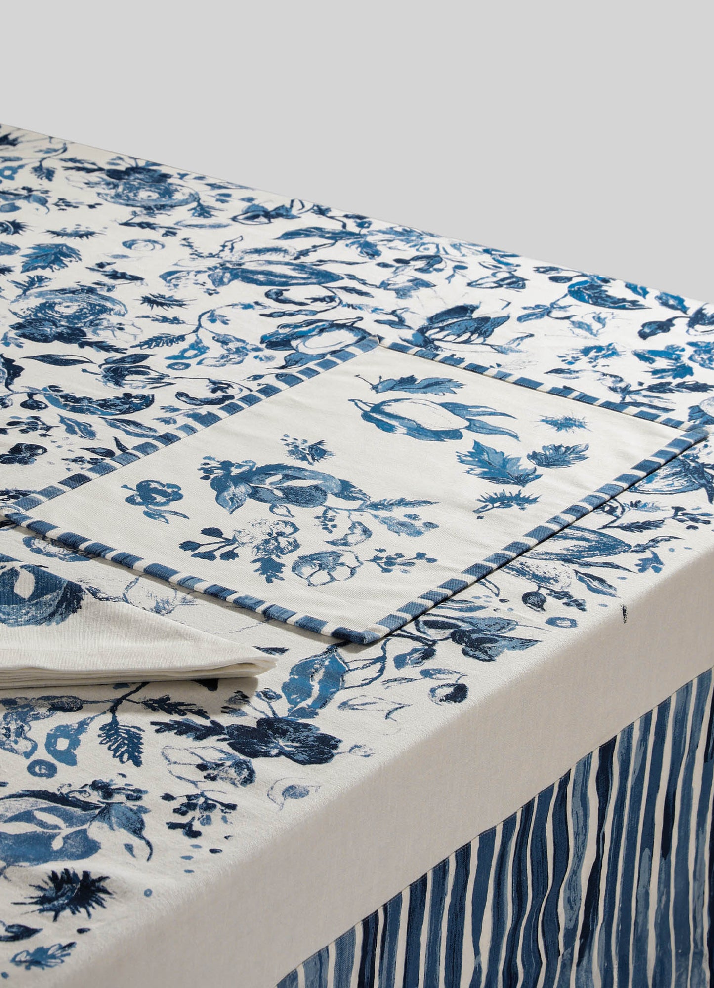 A tablecloth with a blue and white floral pattern and striped borders, made of cotton or linen fabric. The design features large, intricate floral motifs and a clean, classic look suitable for casual or traditional table settings.