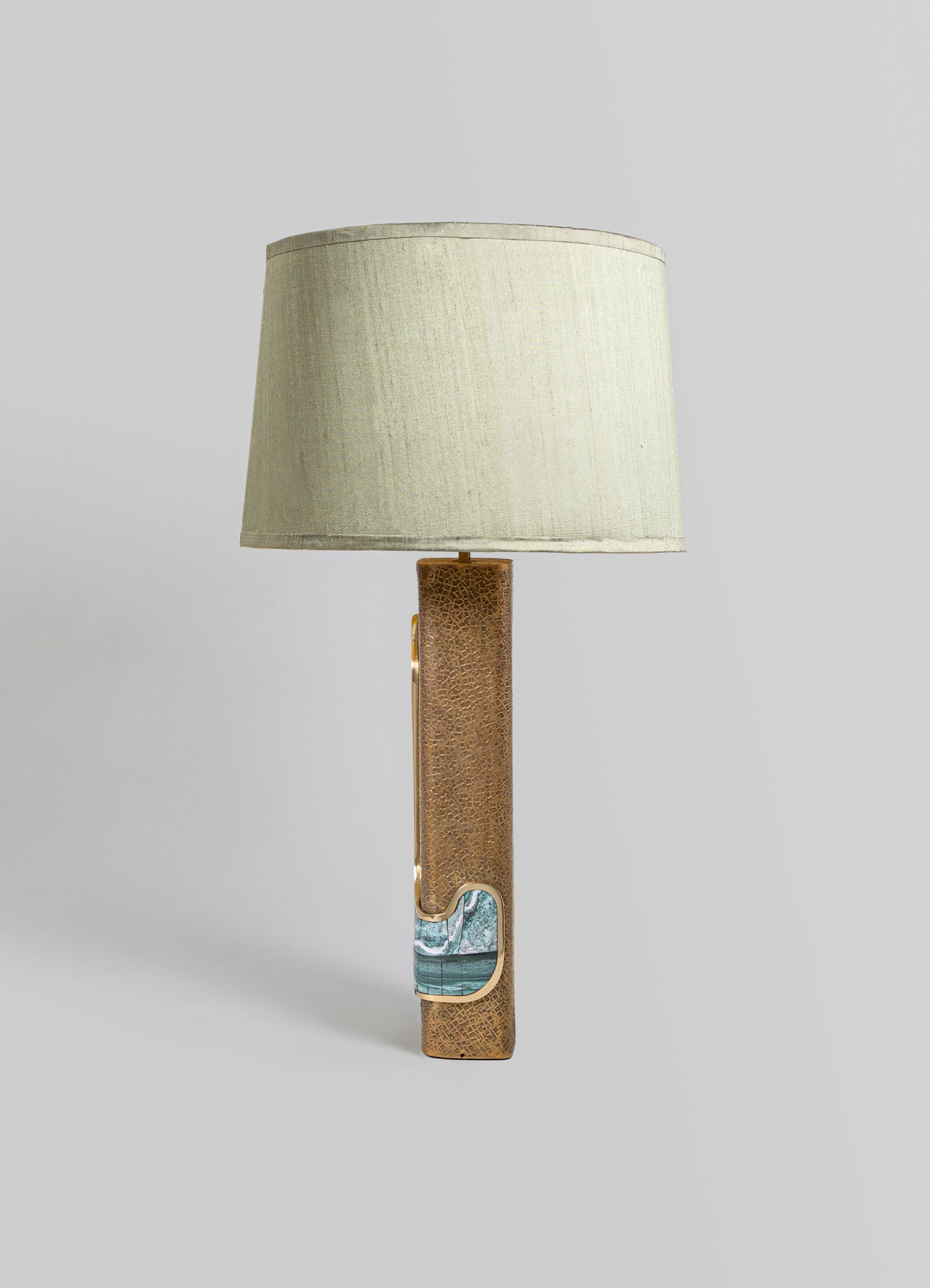 A table lamp with a beige fabric shade and a textured, rectangular base featuring metallic bronze and turquoise accents. The design combines contemporary materials with a minimalist silhouette.