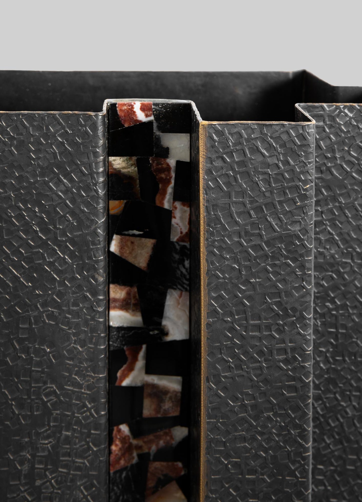 A decorative box with a black textured surface featuring a geometric pattern and a vertical inlay of mosaic stone in warm tones, with a slim metallic gold edge.