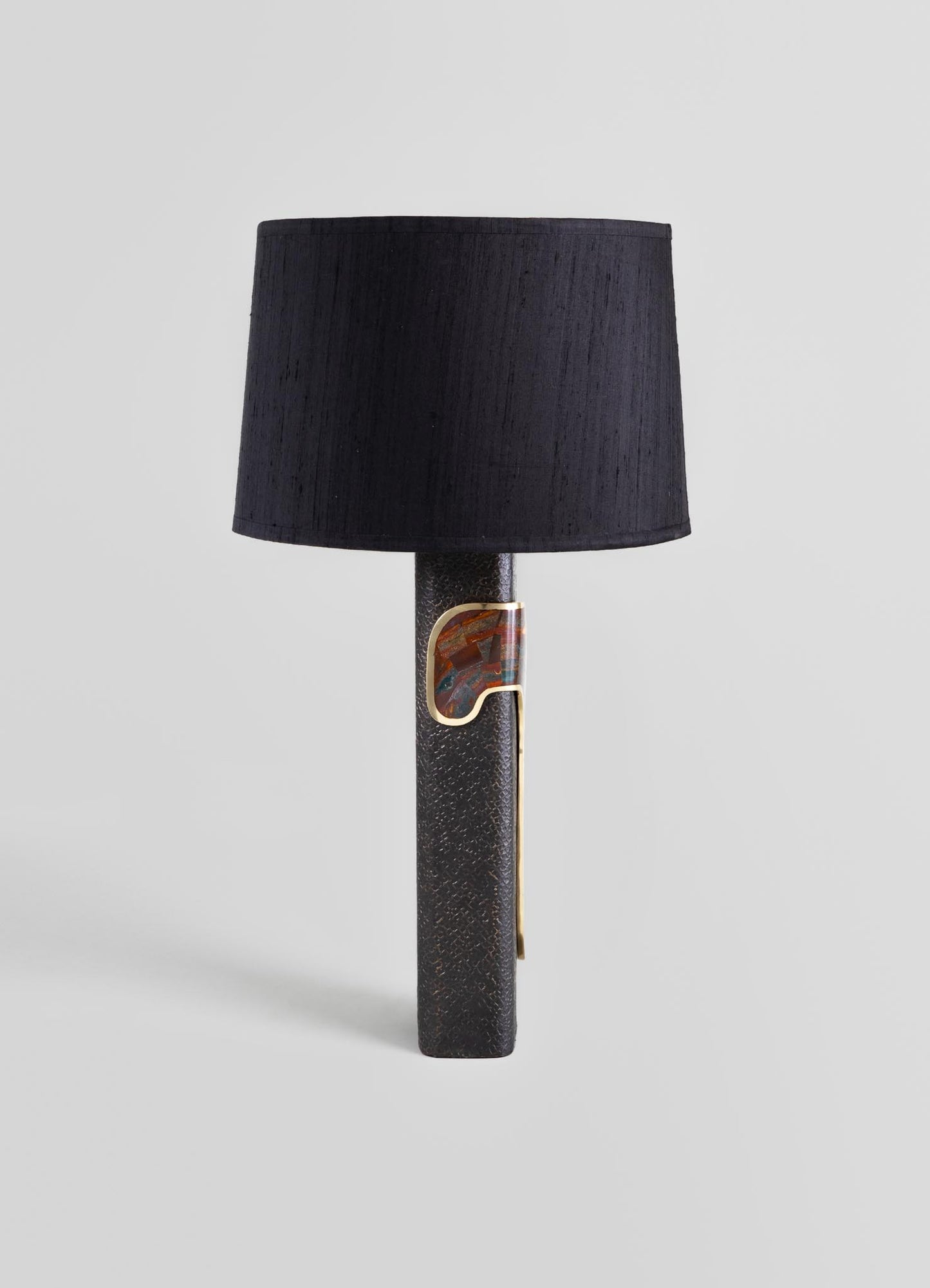 A table lamp with a textured black ceramic base featuring a metal accent in gold and multicolored details, topped with a black fabric lampshade.