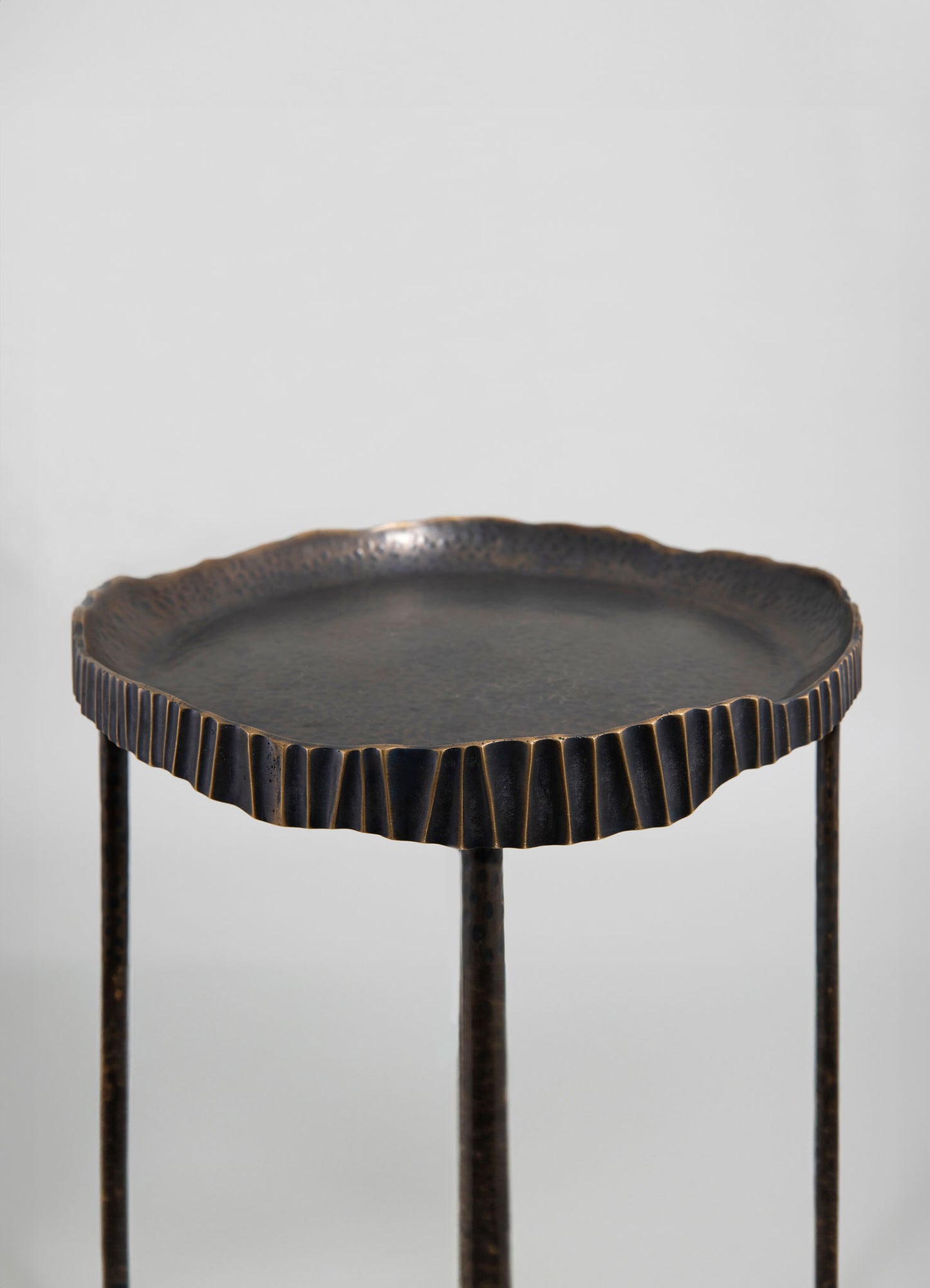 A round, black metal side table with a textured, scalloped edge top and three slender legs, offering a rustic and artisanal style.