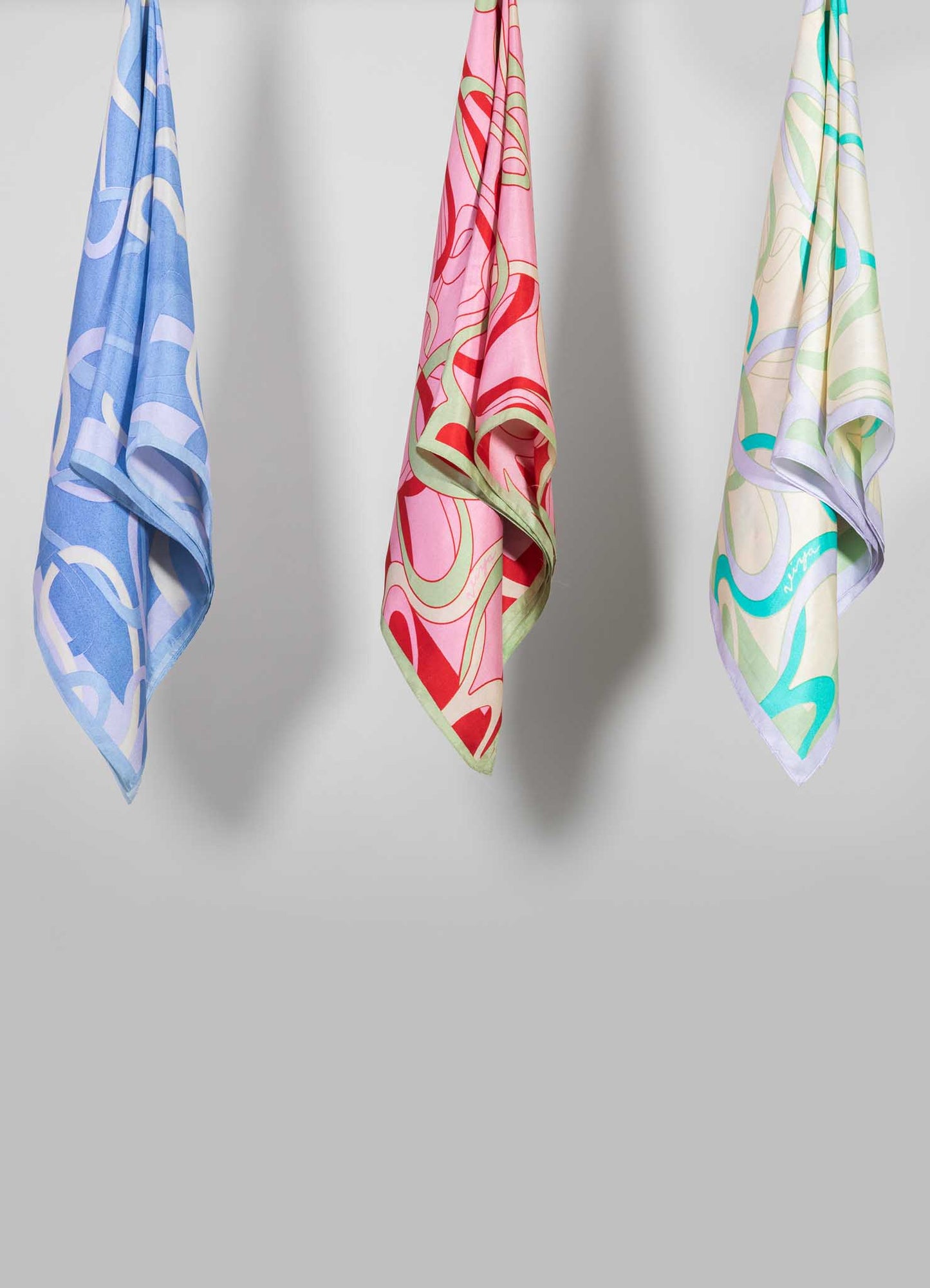 Silk scarves with bold, abstract floral and geometric patterns in pastel and vibrant colors, hanging against a plain background. The scarves feature soft, smooth textures and light, airy designs.