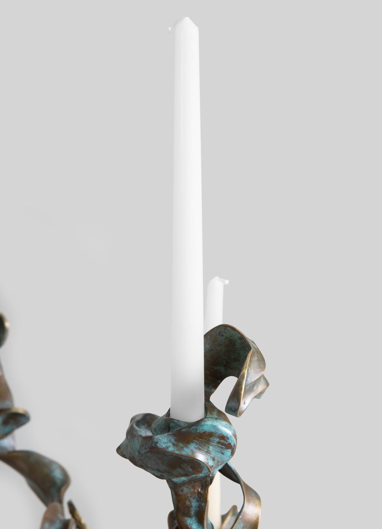 A decorative metal candle holder with an organic, sculptural design featuring a patinated copper finish. It holds tall white taper candles and has a rustic, artistic style with flowing, natural lines.