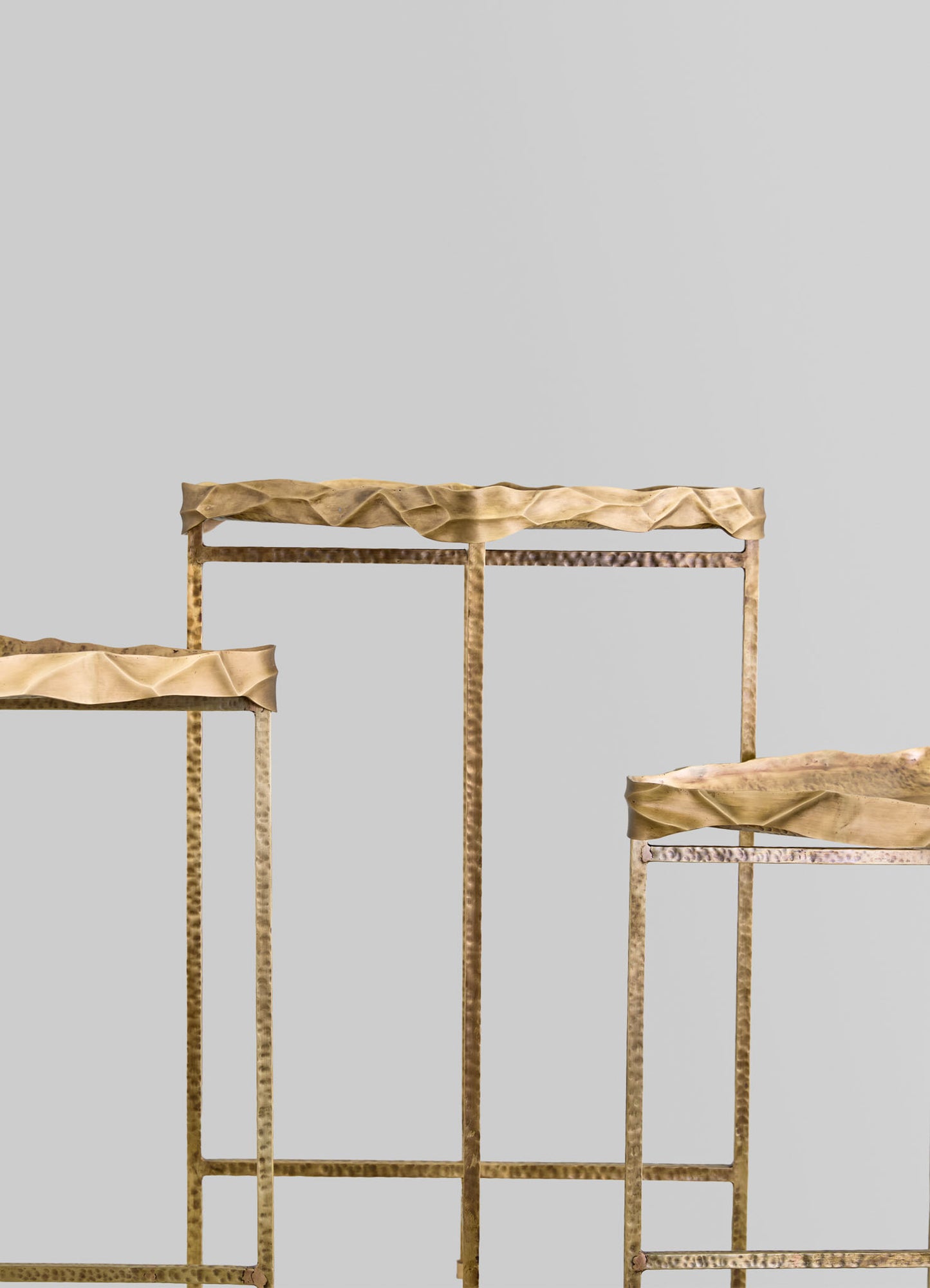 Three metallic gold furniture pieces with hammered and textured finishes, featuring slim legs and wavy, irregular edges on the tabletops. The set conveys a contemporary and artisanal style against a plain, light gray background.