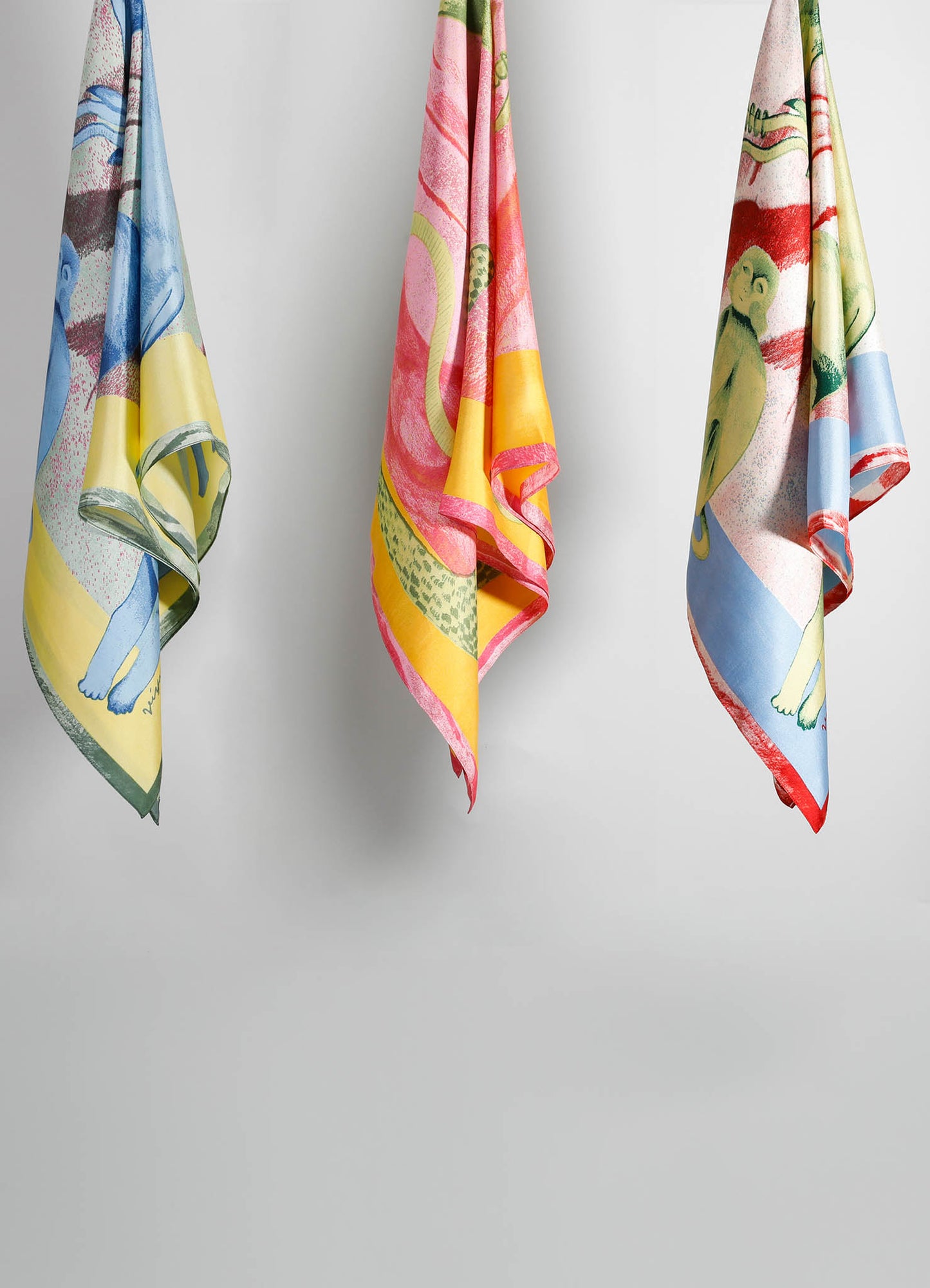 Set of three colorful silk scarves featuring playful animal and abstract motifs in pink, blue, yellow, and green tones, displayed against a plain background. The scarves have a vibrant, contemporary style with detailed printed patterns.