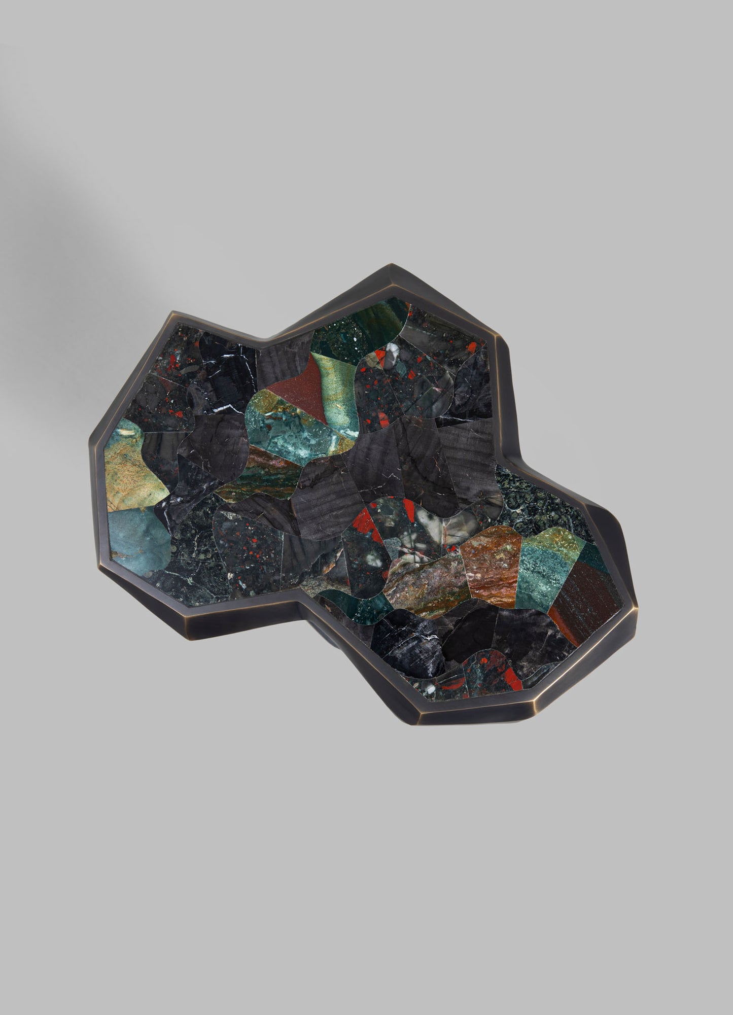 A decorative tray with an irregular, geometric shape featuring a black frame and a surface made of multicolored marble or stone fragments in shades of green, red, black, and beige, arranged in an abstract mosaic pattern.