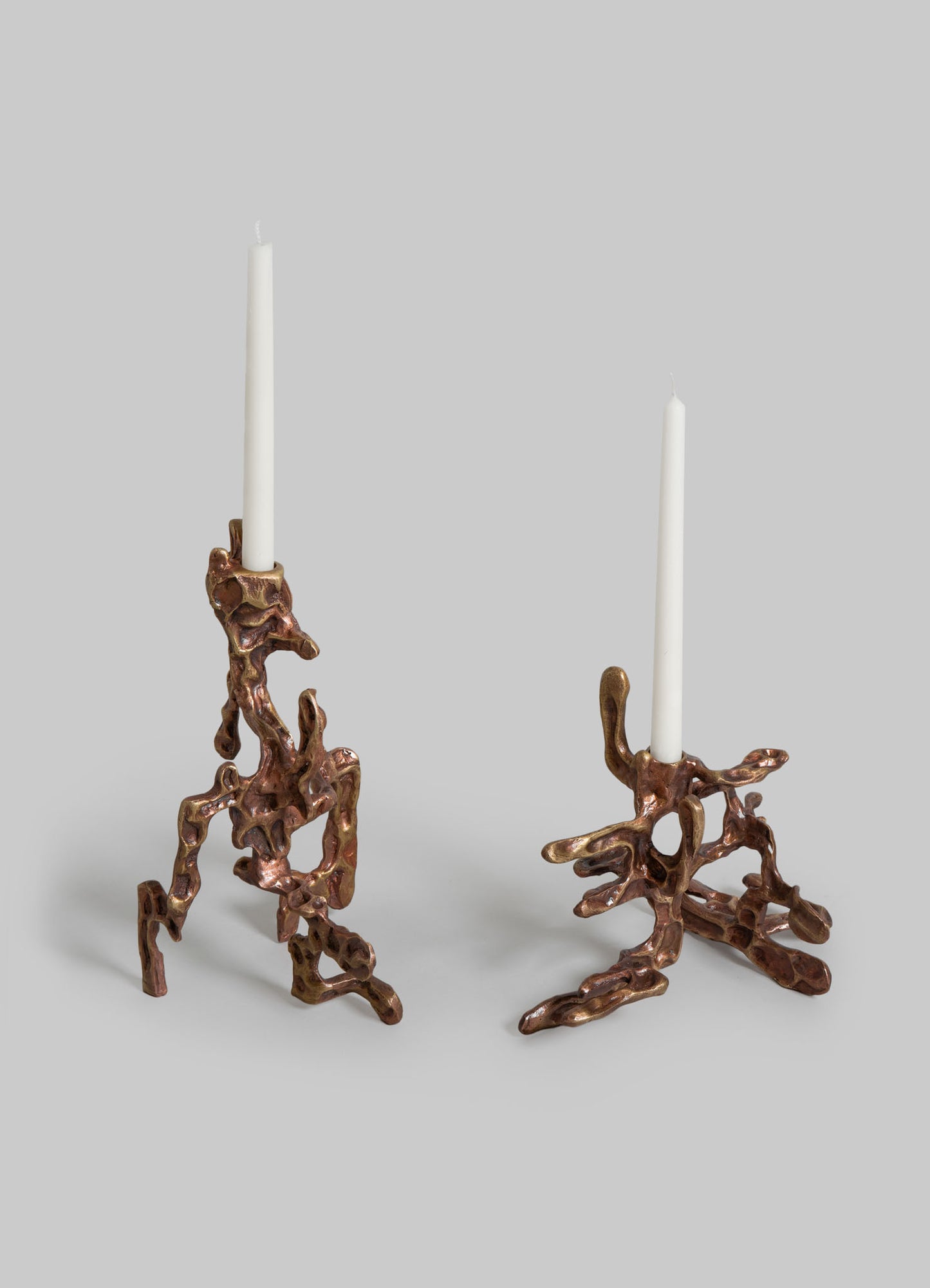 Pair of sculptural candle holders made of textured, organic-shaped metal with a copper finish, each holding a tall white taper candle.