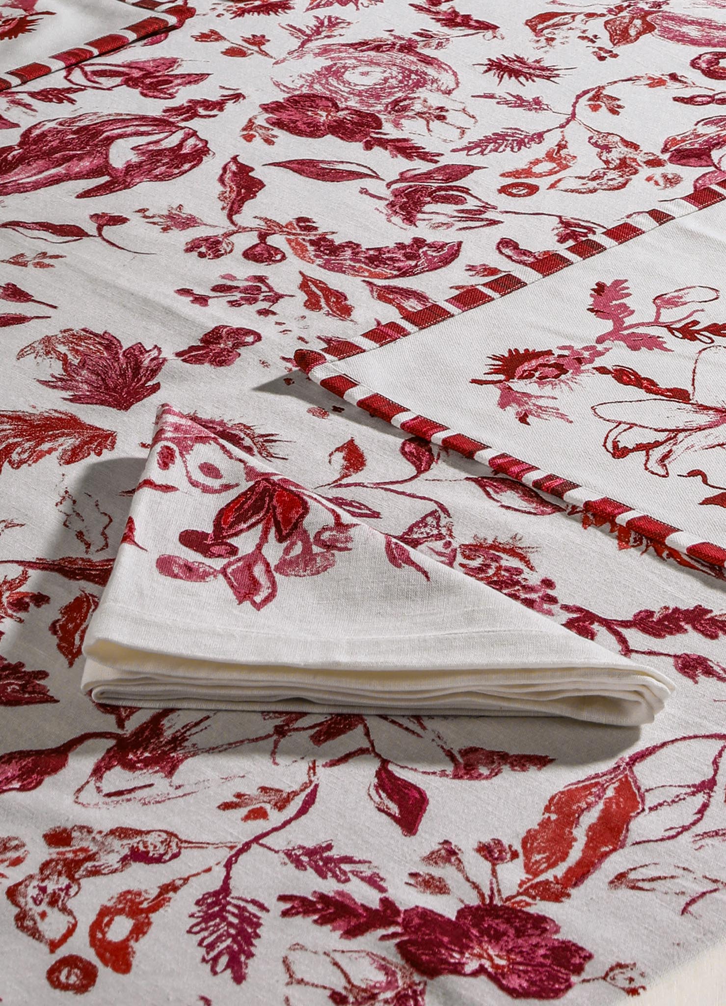 Printed linen tablecloth with a botanical floral pattern in shades of red and pink on a white background, finished with a contrasting red and white striped border. The fabric appears lightweight and smooth, suitable for casual or rustic table settings.