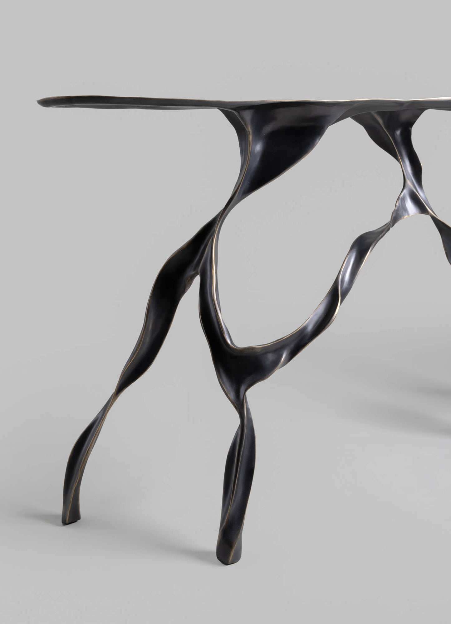 A sculptural table with an abstract, organic design featuring flowing, twisted legs and a smooth, flat surface made of dark, polished metal. The piece has a contemporary style with a sleek, artistic form.