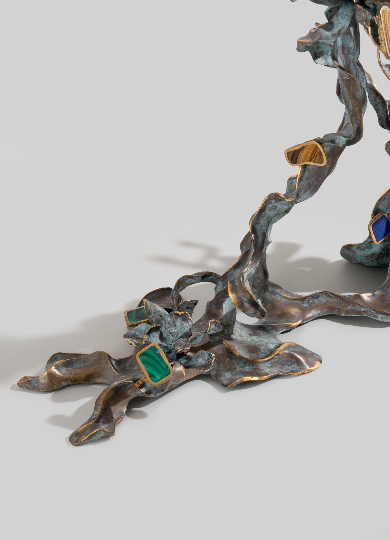 A decorative sculpture made of weathered bronze with flowing, organic shapes accented by rectangular stones in green, blue, and beige tones set in metal frames.