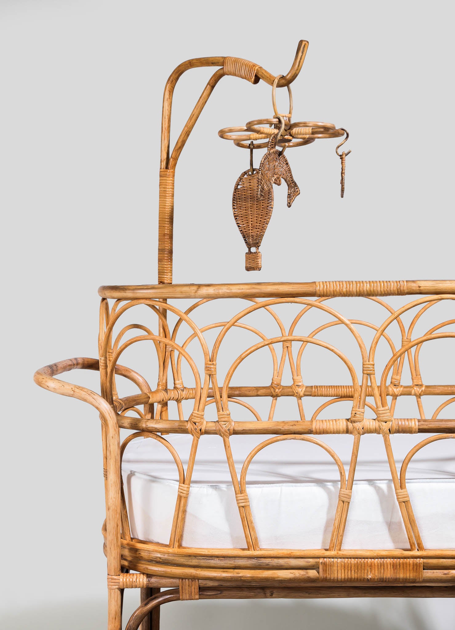 Rattan crib with woven arch-patterned sides and a white fabric lined mattress, featuring a round headboard and curved design in a natural wood finish. A matching mobile with woven shapes hangs above the crib, enhancing a rustic and handcrafted style.