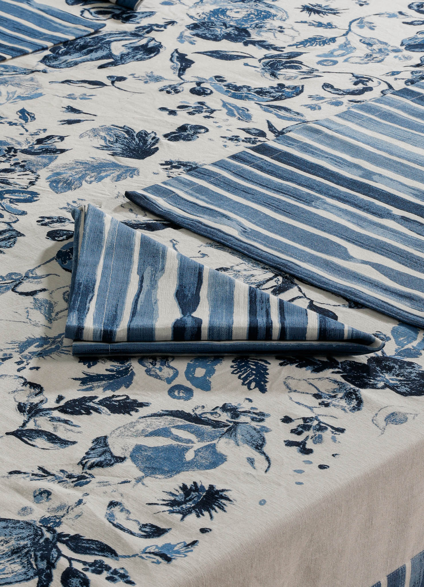 Blue and white printed textile featuring floral and striped patterns, likely made of cotton or linen, with a combination of floral motifs and horizontal stripe designs suggesting table linen or upholstery fabric. The color palette includes various shades of blue on a neutral background, with a contemporary and casual style.