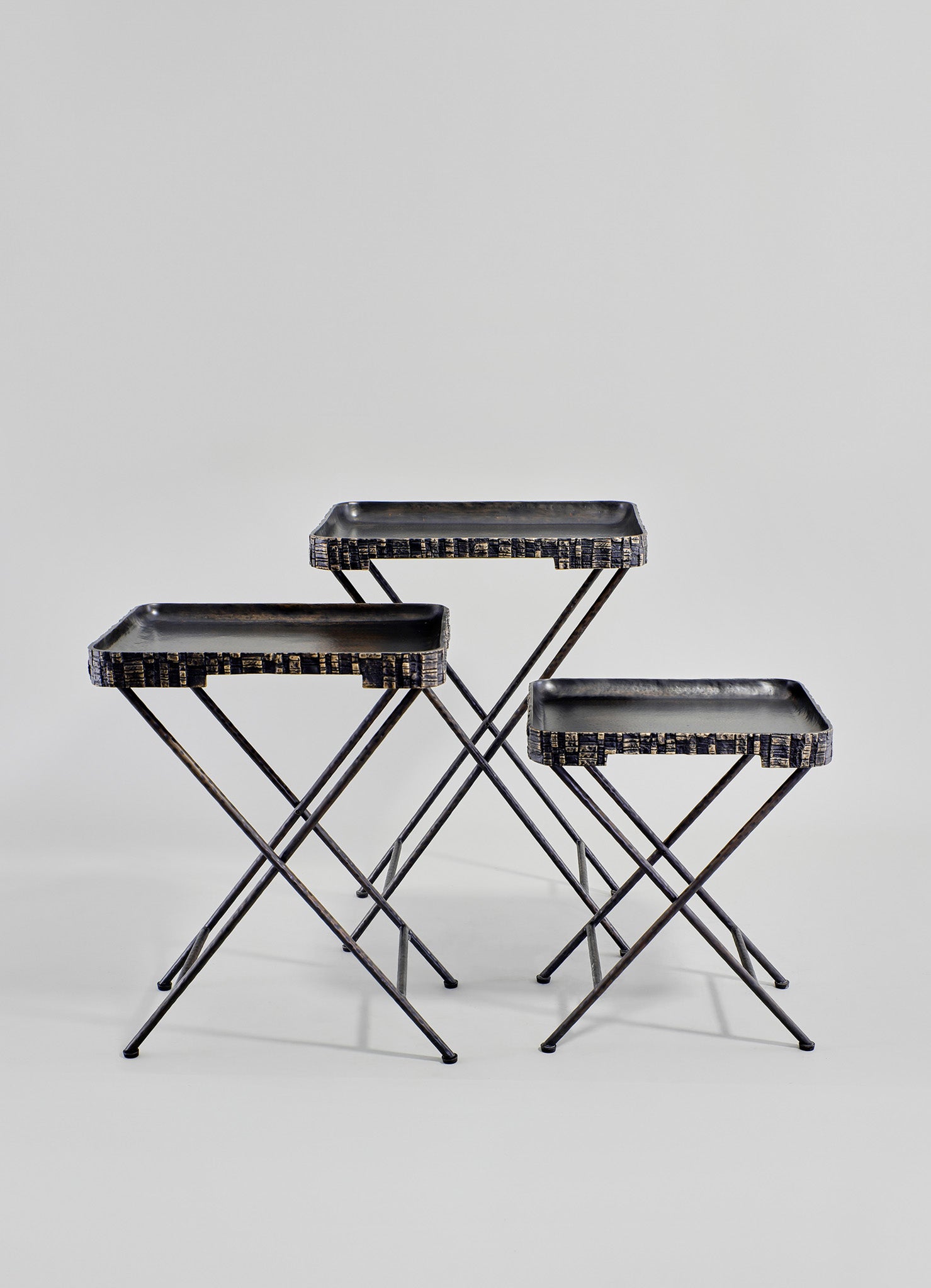 Set of three nesting trays with rectangular blackened metal surfaces and slim, intersecting metal legs, featuring textured wood-like borders in a rustic style.