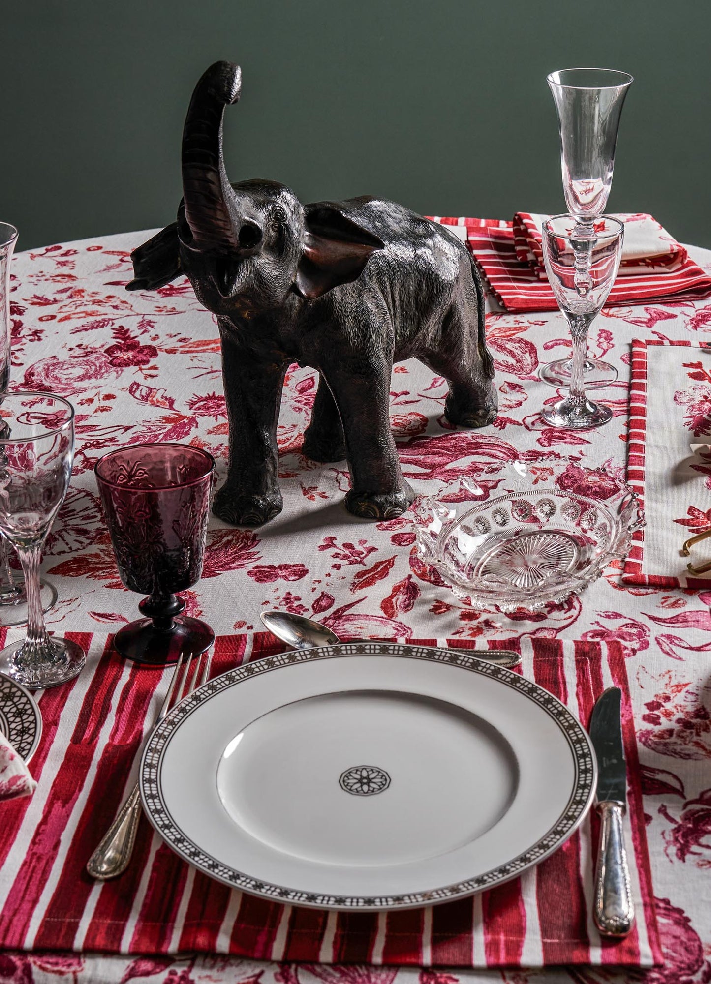 Decorative black elephant sculpture on a table with red and white floral tablecloth, surrounded by clear and purple glassware, porcelain plates with intricate borders, and polished silver utensils. The setting features classic and ornate dinnerware with a mix of transparent and colored glass elements.