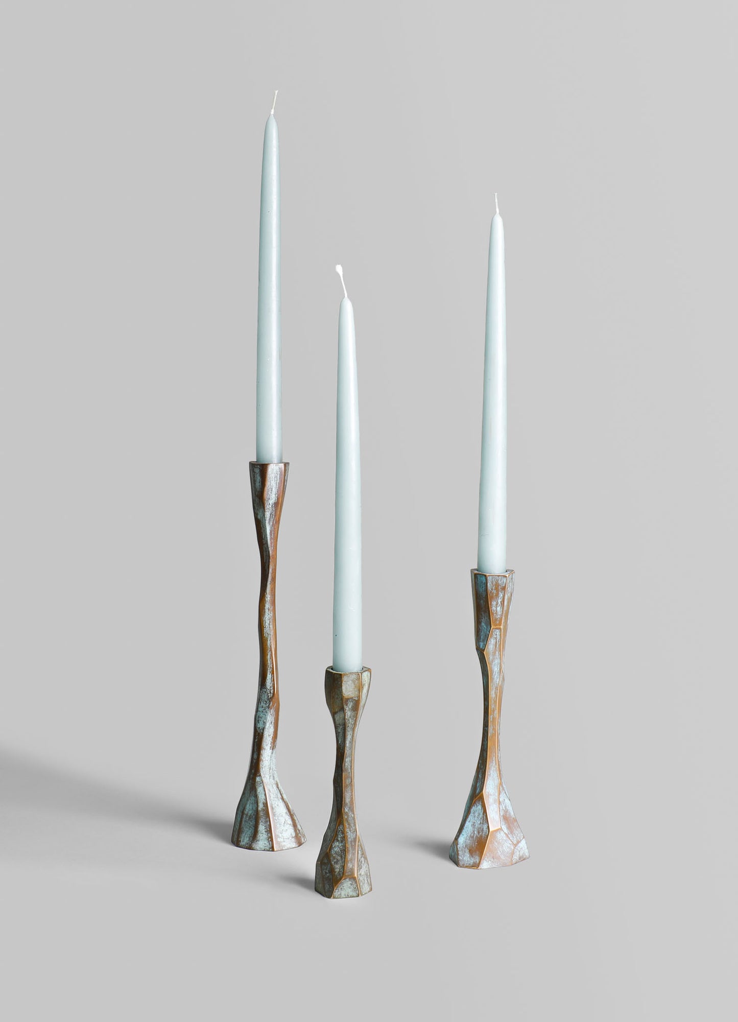 Three taper candles with light blue wax are placed in sculptural metal candleholders with a distressed, oxidized finish, set against a plain, neutral background.