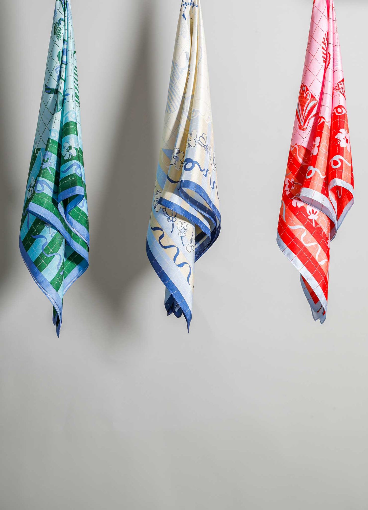 Three patterned tea towels made of cotton or linen, each with a floral motif and geometric border, in colors of green and blue, cream and blue, and red and white, hanging against a plain background.