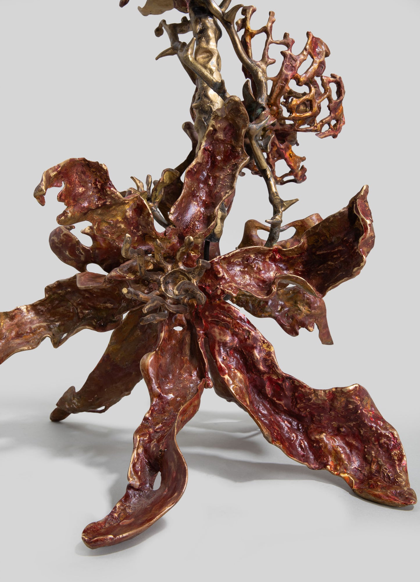 A sculptural art piece featuring intertwined organic forms in metallic gold and reddish-brown finishes, resembling abstract flower or plant motifs. The sculpture emphasizes a rustic, handcrafted style with textured surfaces and irregular shapes.