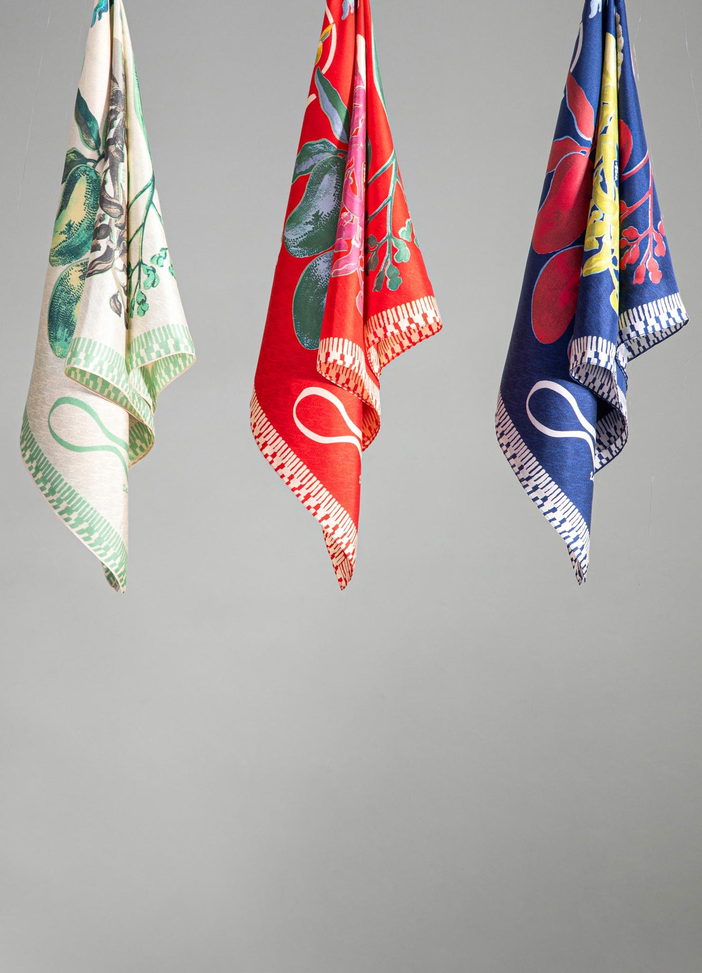 Three hanging fabric towels with floral motifs and bold color schemes: one in cream with green and gray accents, one in red with green and pink accents, and one in navy blue with yellow and pink accents. The towels feature printed patterns with leafy and floral designs and striped borders.