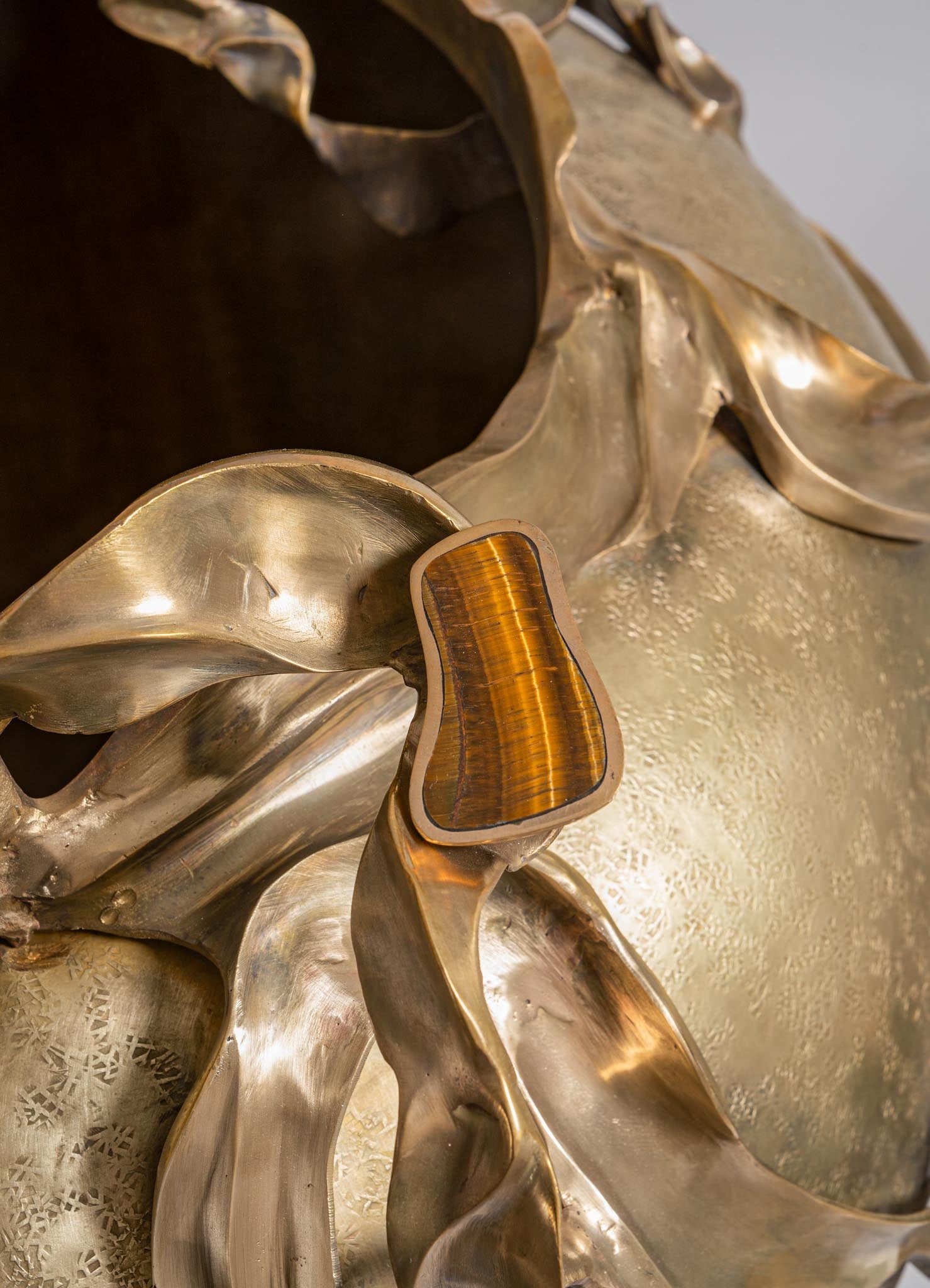 Decorative metal sculpture with flowing, sculptural brass elements and a polished, warm-toned tiger's eye stone inset, featuring a textured bulbous surface with a reflective finish, in an artistic contemporary style.