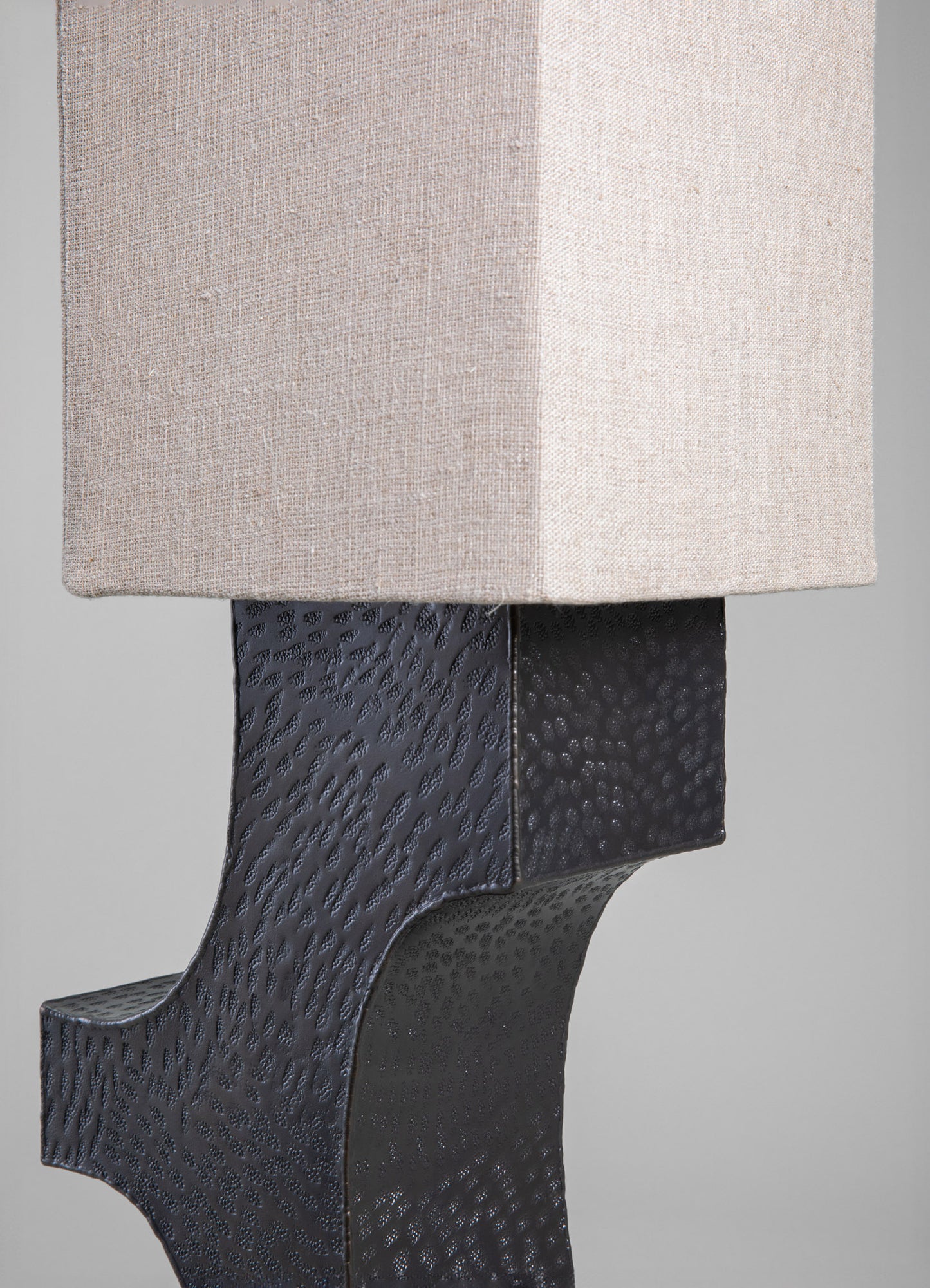 A table lamp with a rectangular fabric shade in neutral beige and a textured black metal base with a hammered finish. The design combines contemporary style with handcrafted metalwork.