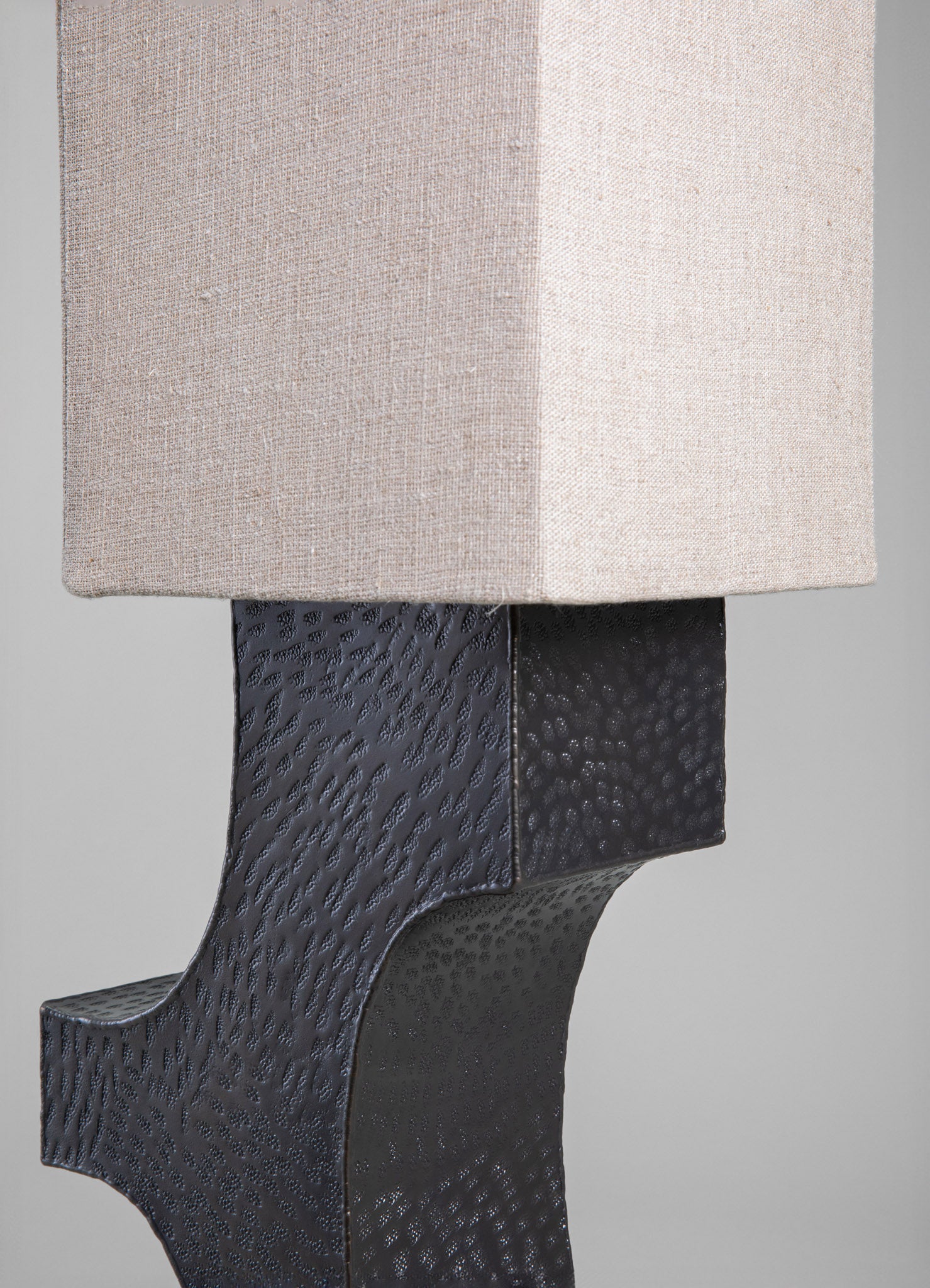 A table lamp with a rectangular fabric shade in neutral beige and a textured black metal base with a hammered finish. The design combines contemporary style with handcrafted metalwork.