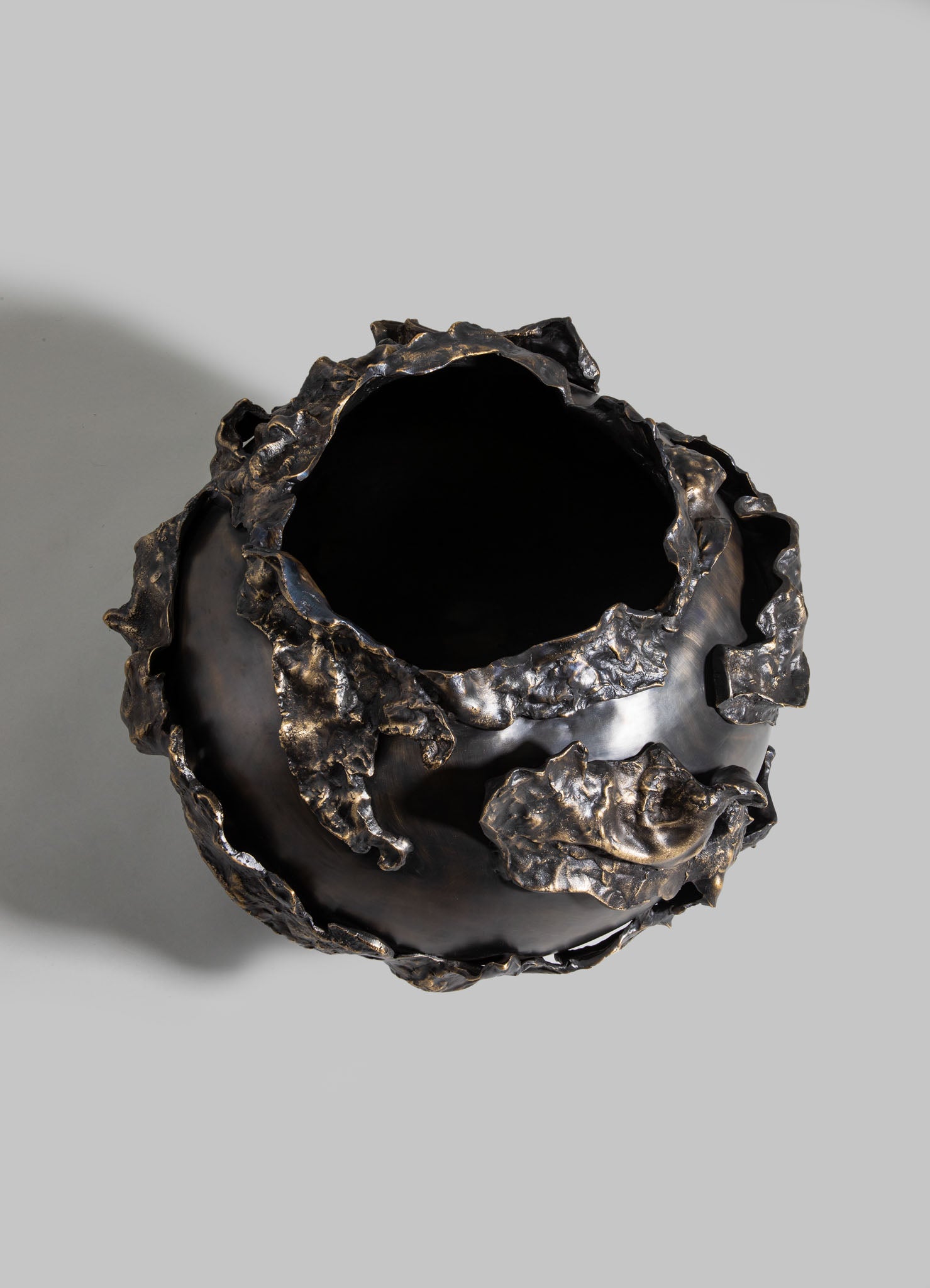 A sculptural vase with an irregular, textured surface in dark metallic tones, resembling crumpled or molten metal, with an open top and organic, sculptural form.