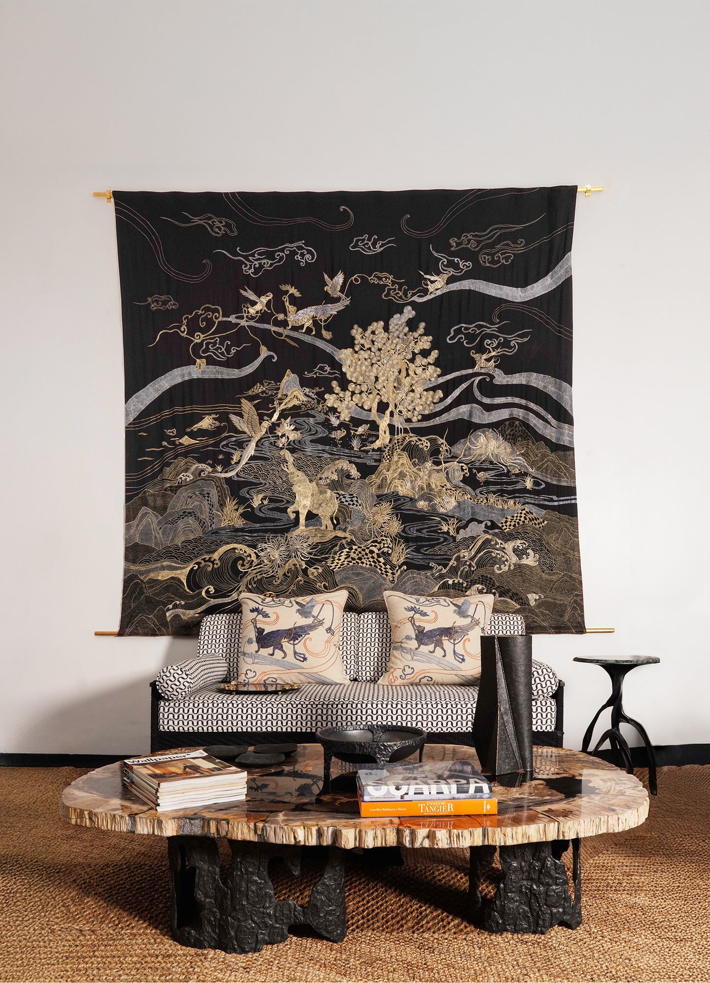 A living room with Viya's Dreamscape Black & Gold Embroidered Bedcover as a tapestry, a patterned sofa with two pillows, and a round wooden coffee table holding books and a black vase, all set on a woven rug.