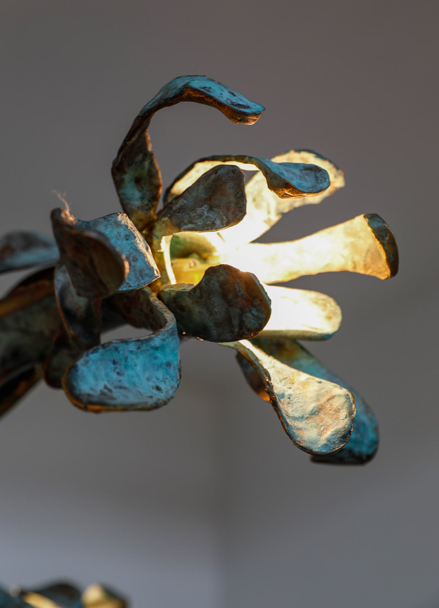 A decorative metal sculpture resembling a flower with textured, weathered petals in shades of blue, brown, and gold. The piece has an organic, handcrafted style with a warm glow emanating from behind the petals, highlighting its intricate details and rustic finish.
