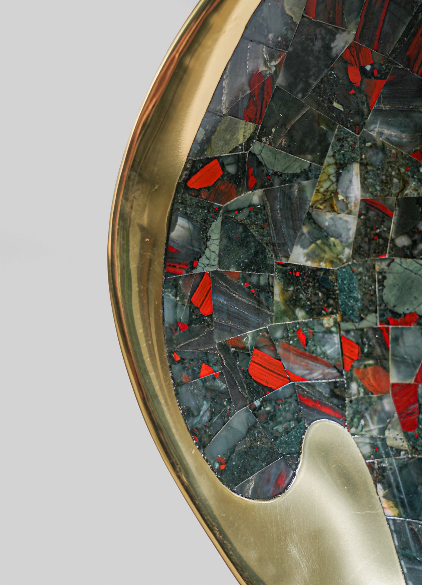 A mosaic tabletop with irregular pieces of dark green, red, and gray stone, encased in a polished brass frame with a smooth, curved edge, presenting a contemporary design with bold, contrasting colors and textures.