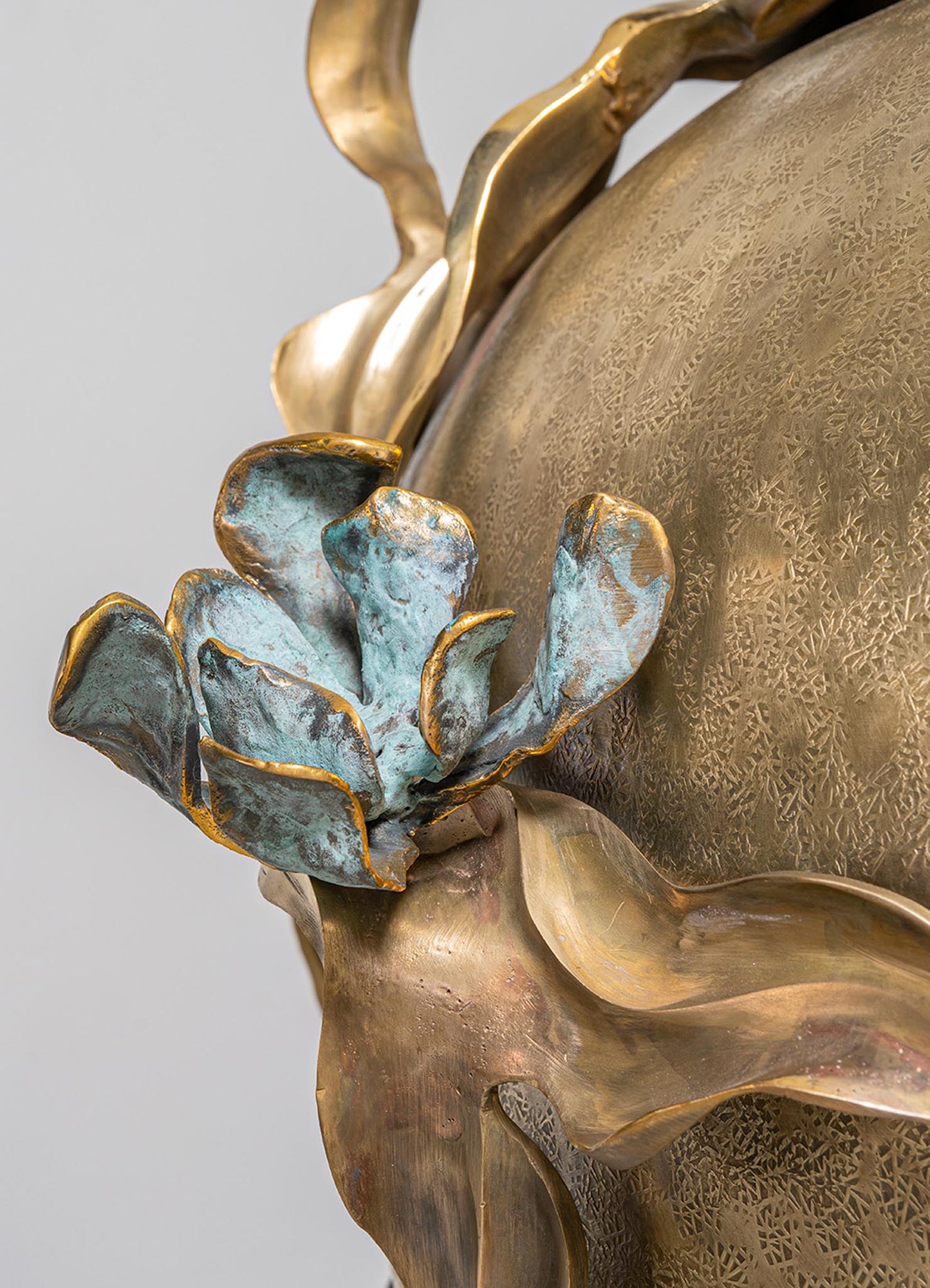 Decorative metal sculpture with a floral motif, featuring a textured gold finish and a blue-green patina on the flower petals, mounted on a curved base with an elegant, handcrafted design.