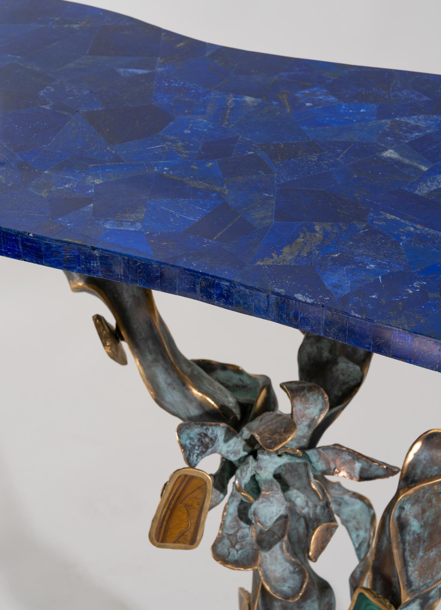 A table with a polished mosaic top made of irregular blue and gold-toned stone tiles, featuring a sculptural metal base with organic, leaf-like forms finished in a metallic bronze tone.
