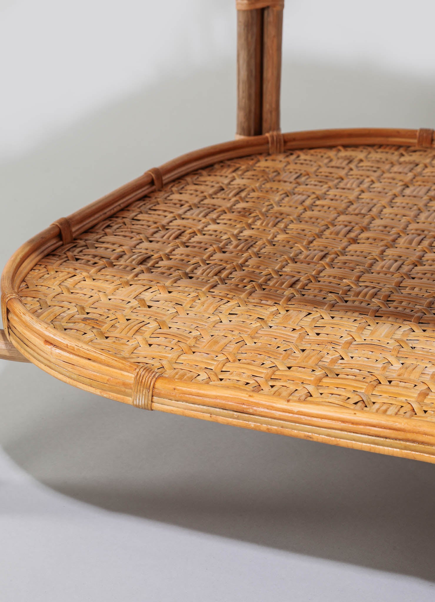 A close-up of a woven rattan tray with a rounded edge and a natural wood frame, showcasing intricate basket-weave patterns and a light tan color. The tray is supported by a wooden stand with vertical legs, emphasizing rustic and handcrafted design elements.