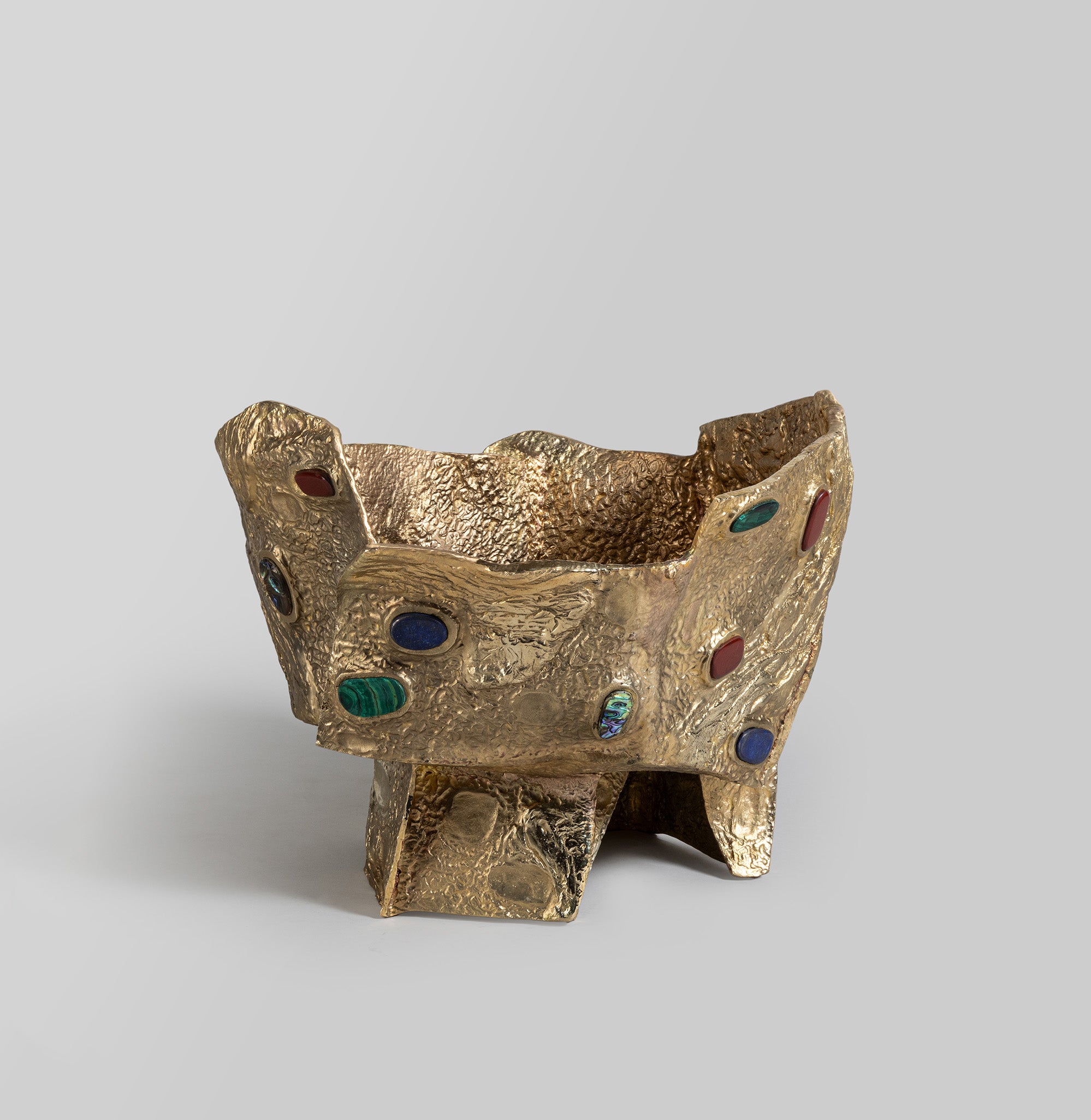 A gold-toned metal bowl with a textured, irregular surface and colorful oval embellishments, supported by three short legs. The design features a handcrafted, rustic style with a tactile finish.