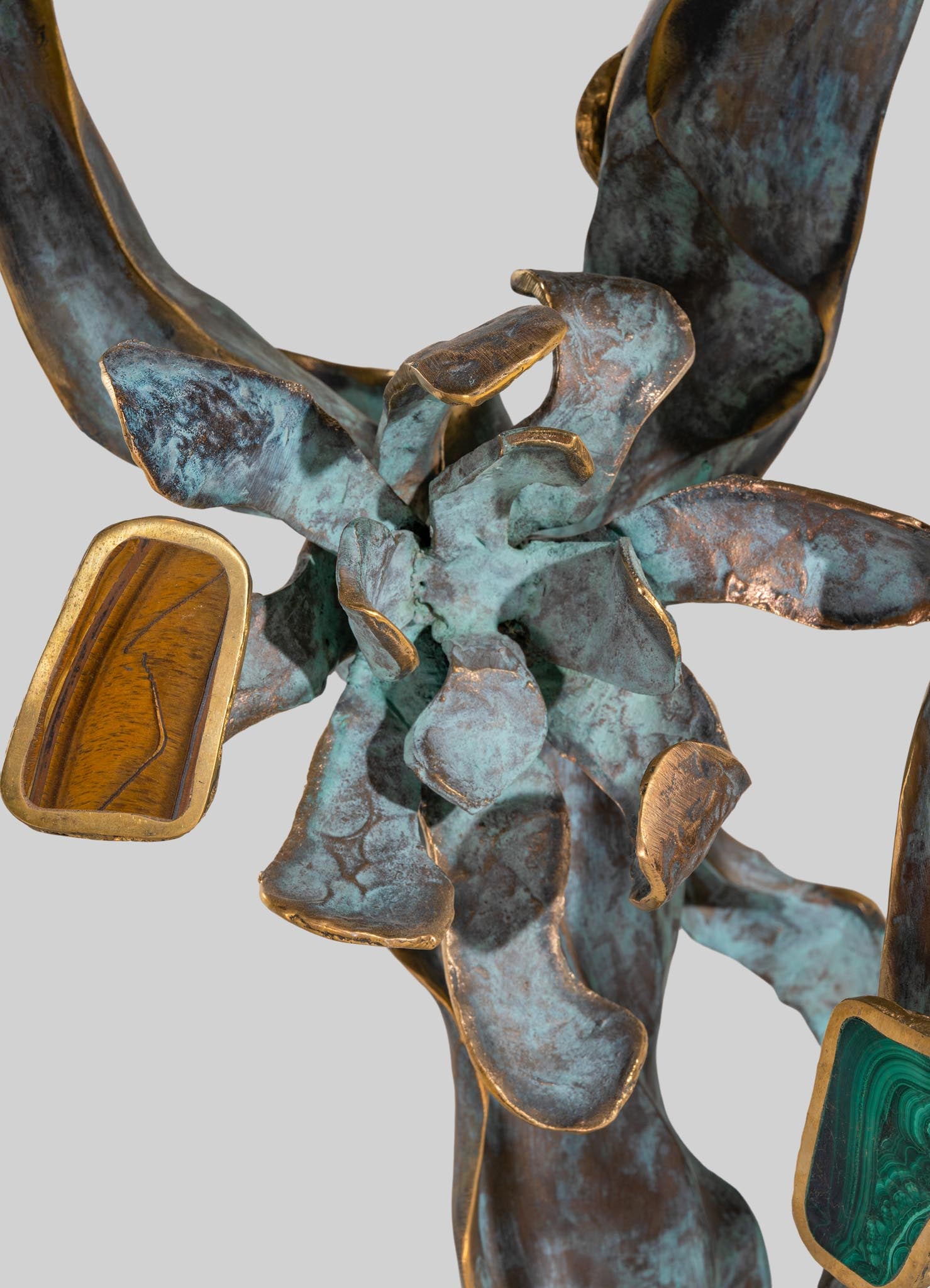 A decorative metal sculpture featuring a floral motif with textured, oxidized blue-green and bronze finishes. The sculpture includes gold-accented leaf or petal shapes and incorporates embedded slices of polished semi-precious stones, such as malachite and tiger's eye.