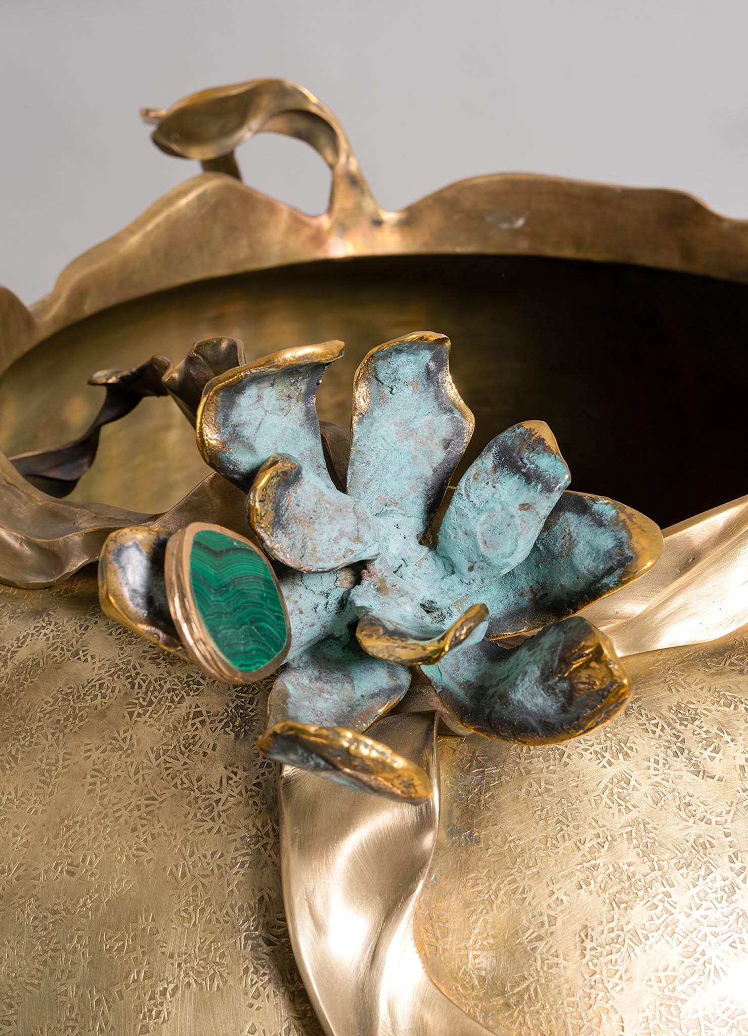 Decorative metal tray with intricate cut-out patterns, featuring a floral-inspired brass sculpture with a patina finish and a green malachite ring with a gold band. The design combines luxury materials with organic motifs, creating a handcrafted artistic display.