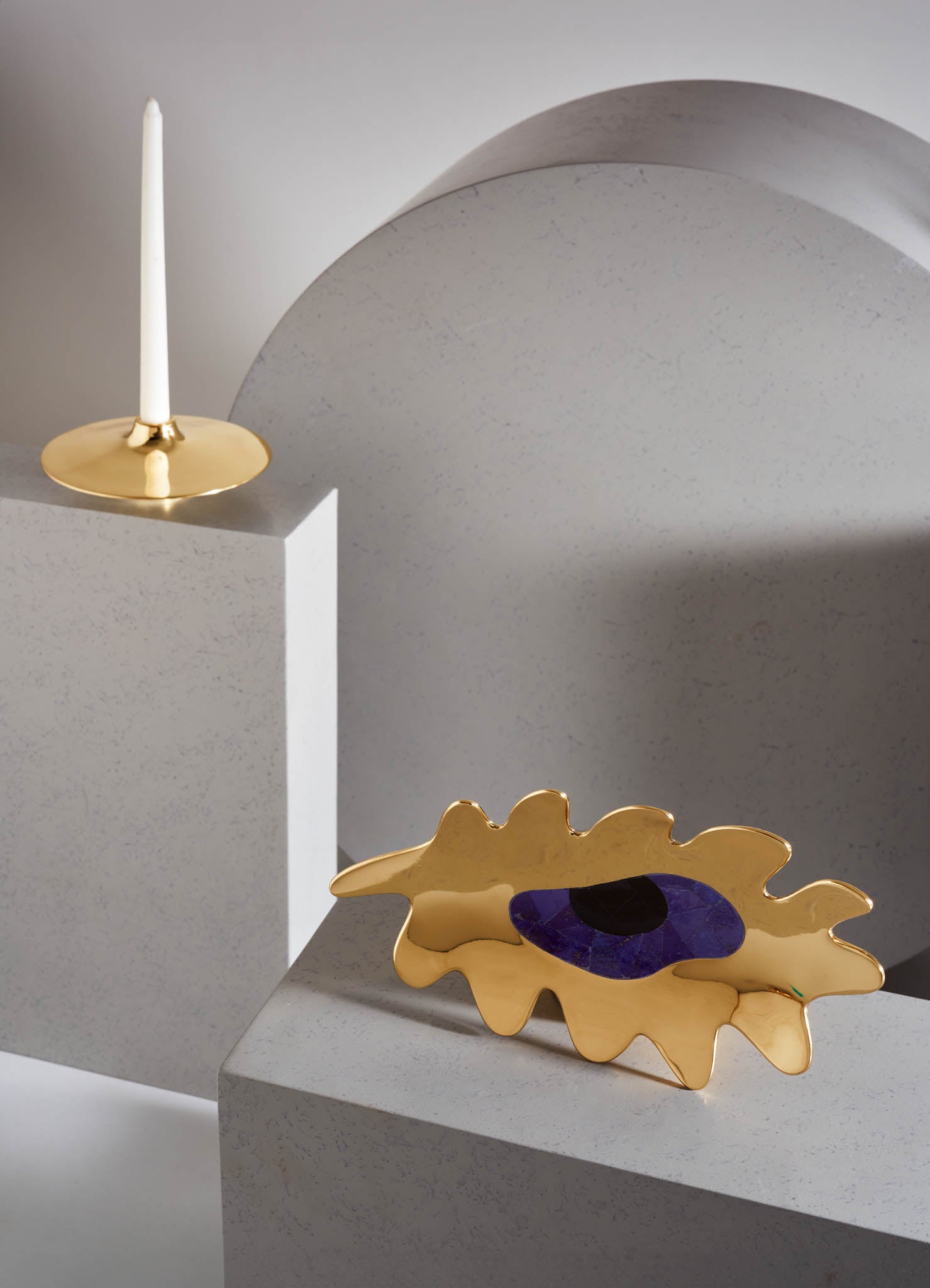 Decorative gold tray with organic, irregular shape and purple inlay, placed on a textured light grey surface. A tall white candle in a gold candlestick is positioned on a nearby grey block, complemented by smooth, rounded curves and minimalist design.
