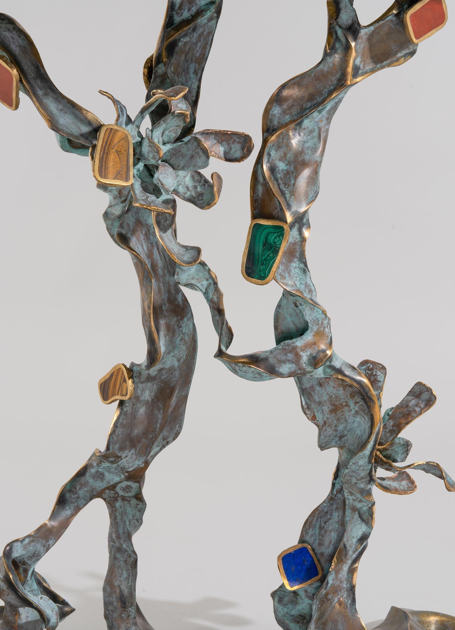 A sculpture resembling a tree branch with a textured, oxidized metal finish in bluish-green and bronze tones, featuring inset rectangular stones of varying colors including green, beige, and blue.
