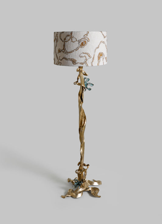Decorative floor lamp with a sculptural, brass-like base resembling a twisted branch and adorned with small, blue-green metallic accents. The drum shade is fabric with embroidered, floral or abstract motifs in neutral tones.